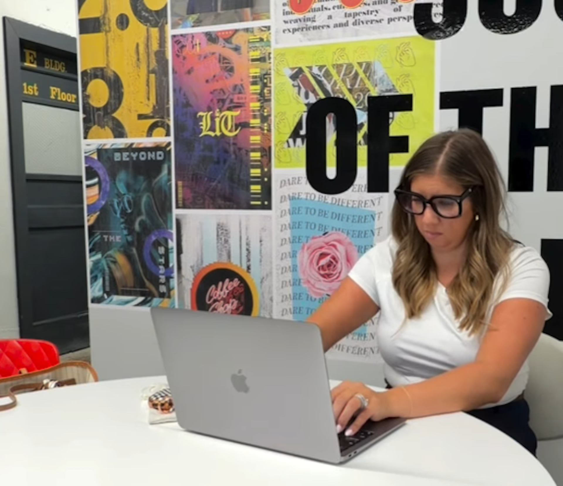 A woman with glasses and long hair working on a laptop at a white table, with a colorful bulletin board behind her displaying various posters and signs.