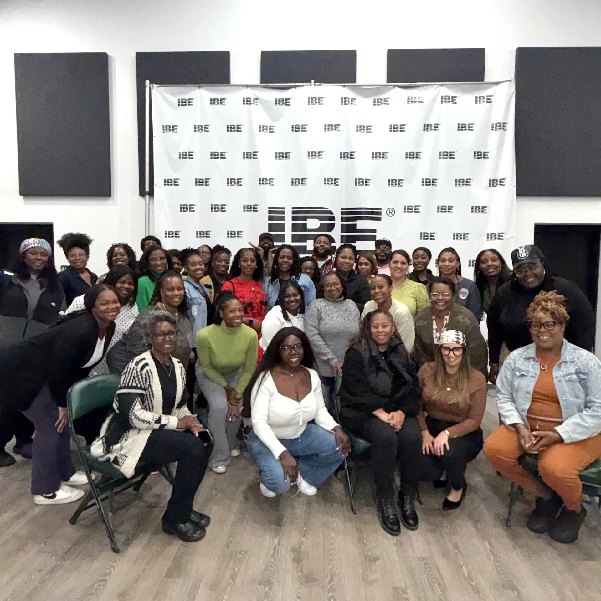 A large group of people gathered together for a group photo in an indoor space with a banner that reads 'IBE' behind them.