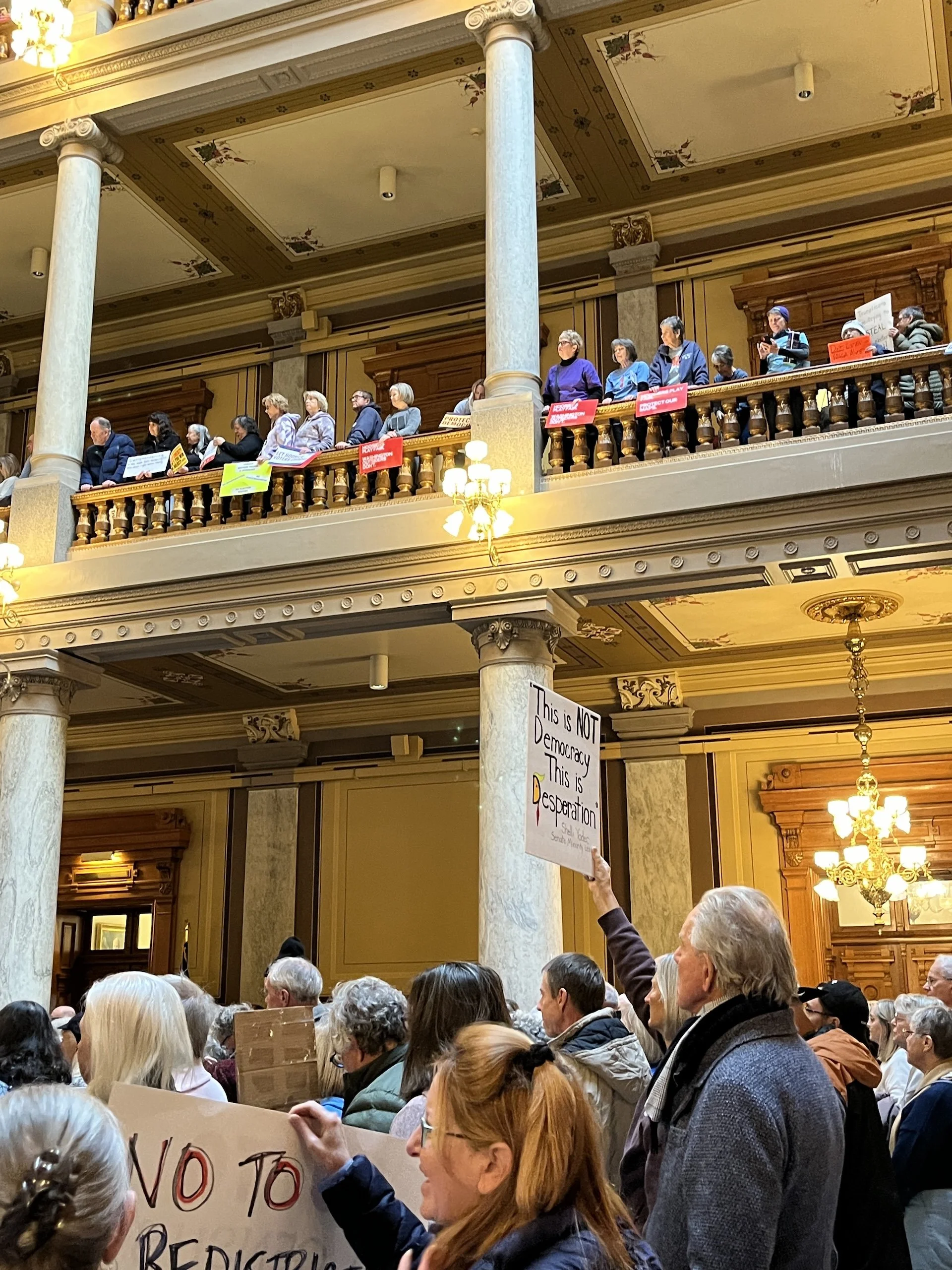 Crowd of people inside a large, ornate building, some holding signs. The building has decorative columns, wood paneling, and chandeliers.