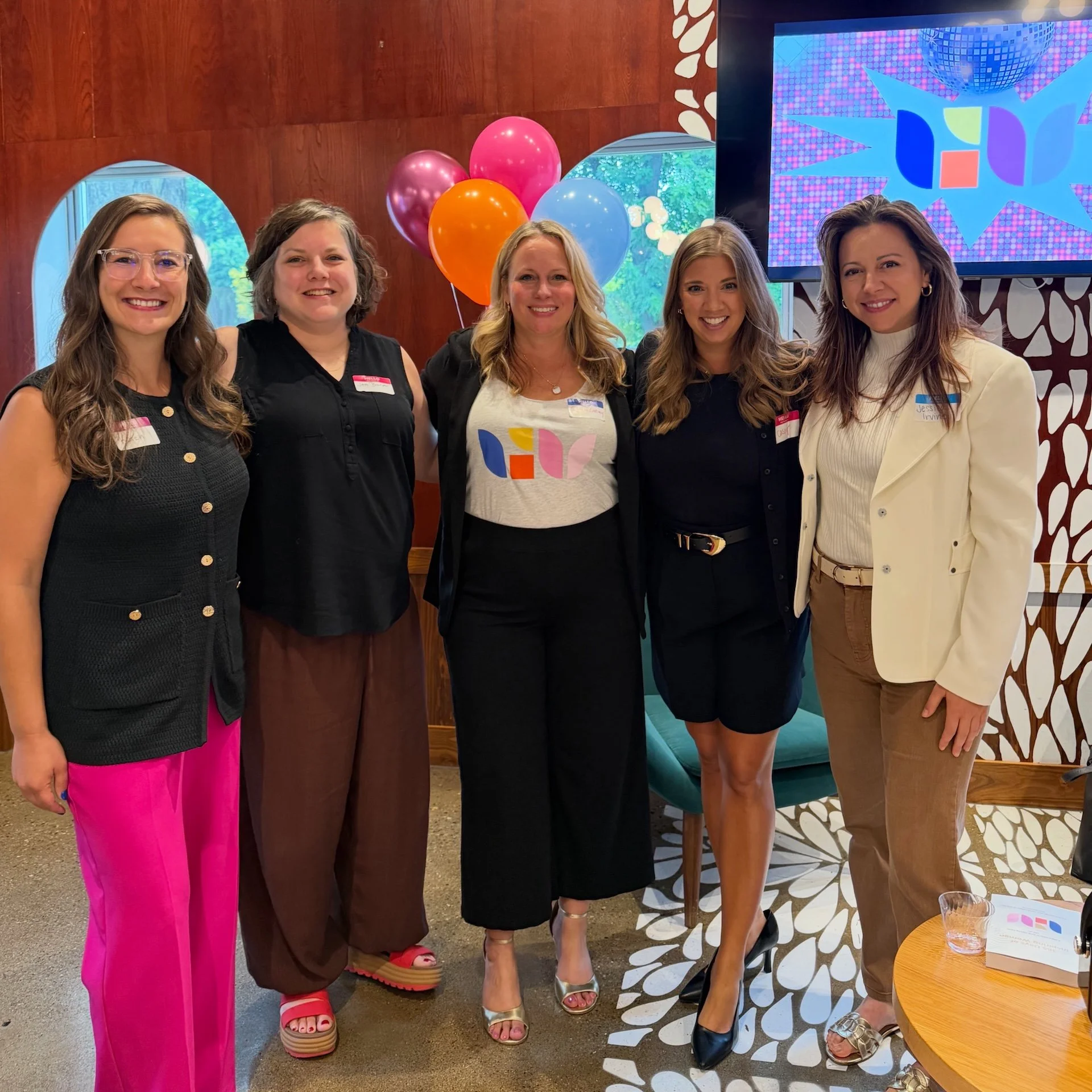 Group of five women smiling and standing together indoors with colorful balloons and decorative background.