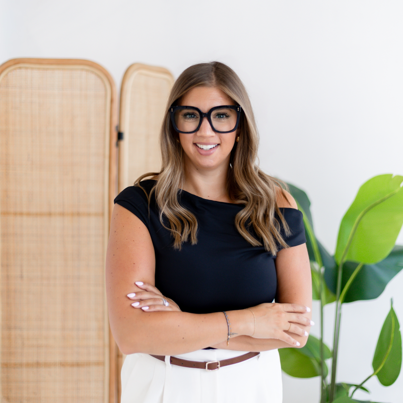 Casey, Polished Founder, smiling while wearing a black top and white pants, standing indoors near a green plant and a rattan room divider.
