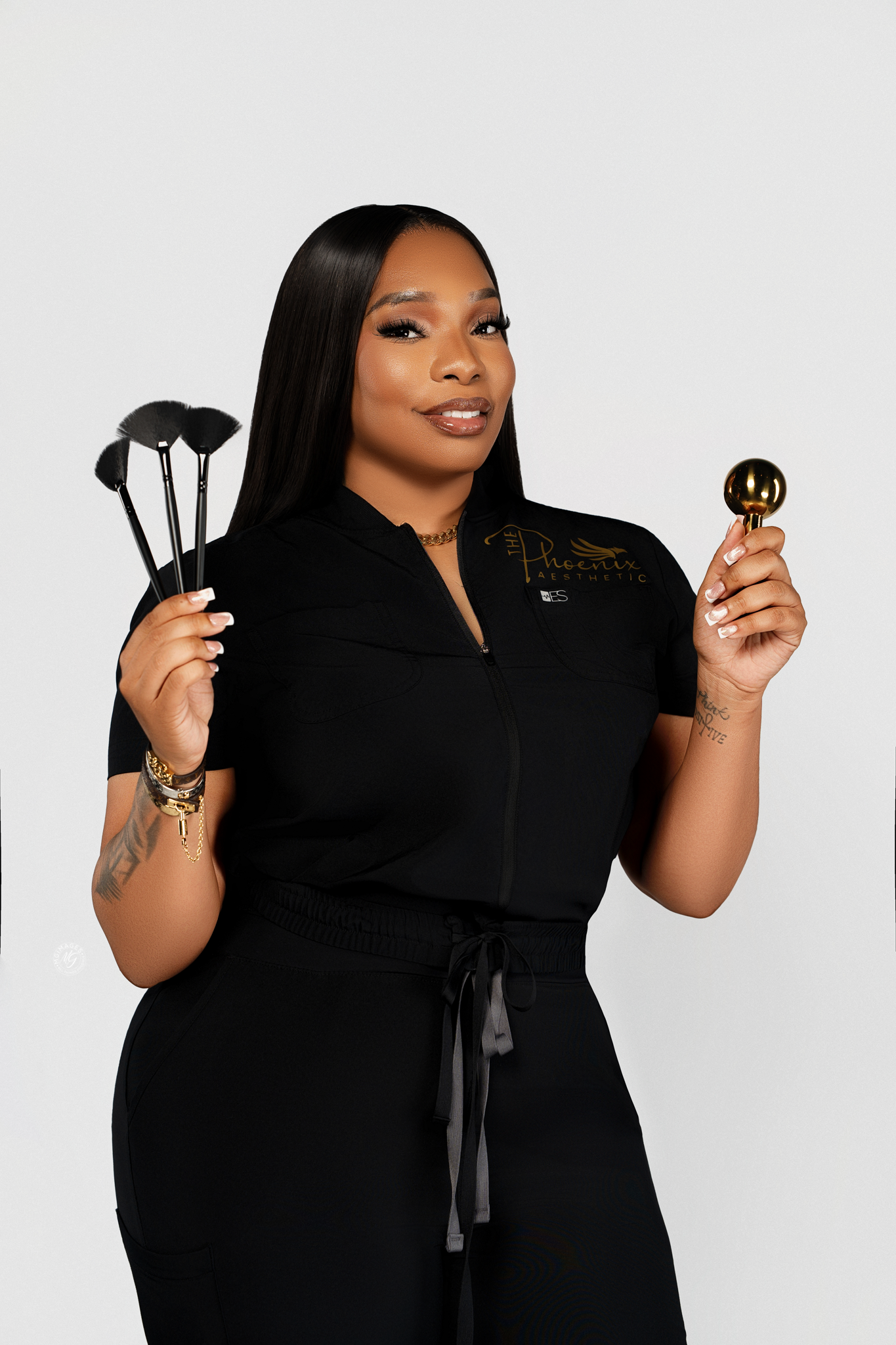 A woman with long black hair, wearing black athletic wear, holding four makeup brushes in one hand and a gold makeup brush in the other, standing against a plain white background.
