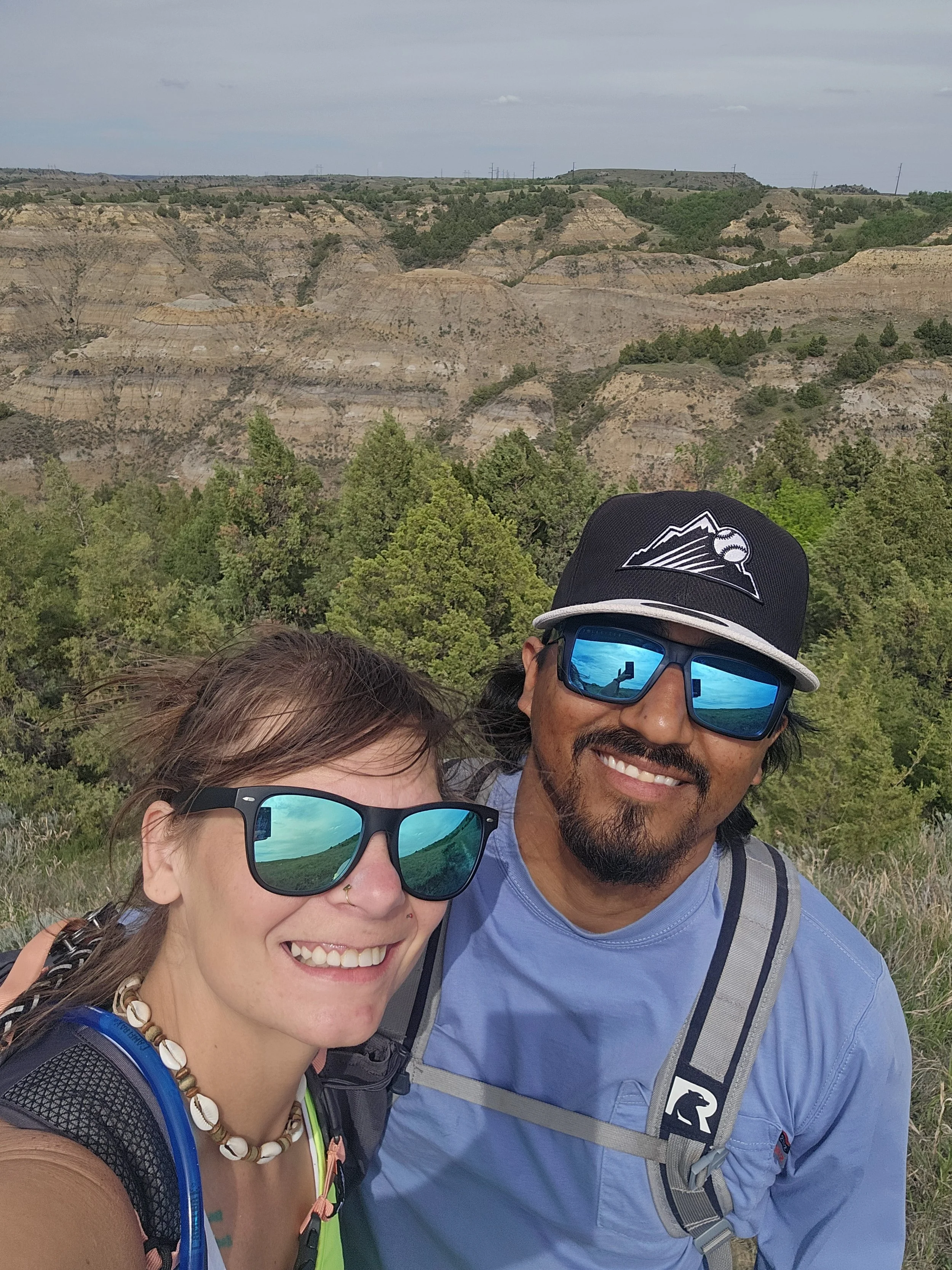A smiling man and woman wearing sunglasses with blue reflective lenses, hiking in a mountainous area with green trees and layered rock formations in the background.
