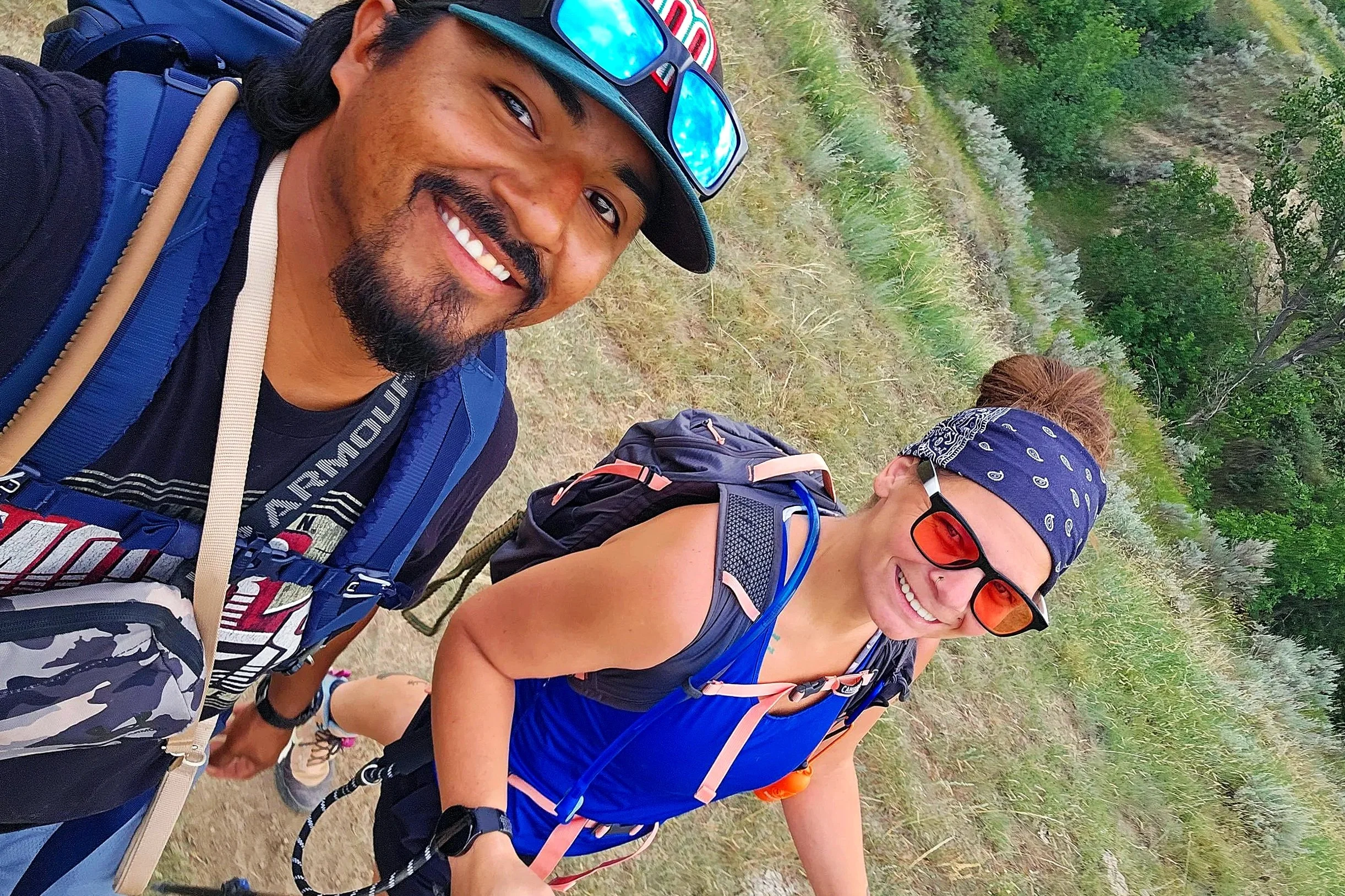 Two smiling women and a smiling man hiking outdoors on a trail with trees and grass in the background. They are wearing athletic clothing, backpacks, and sunglasses.