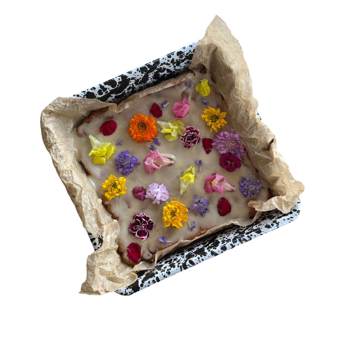 A square dessert topped with white icing and decorated with colorful edible flower petals, served on parchment paper in a black and white patterned box.