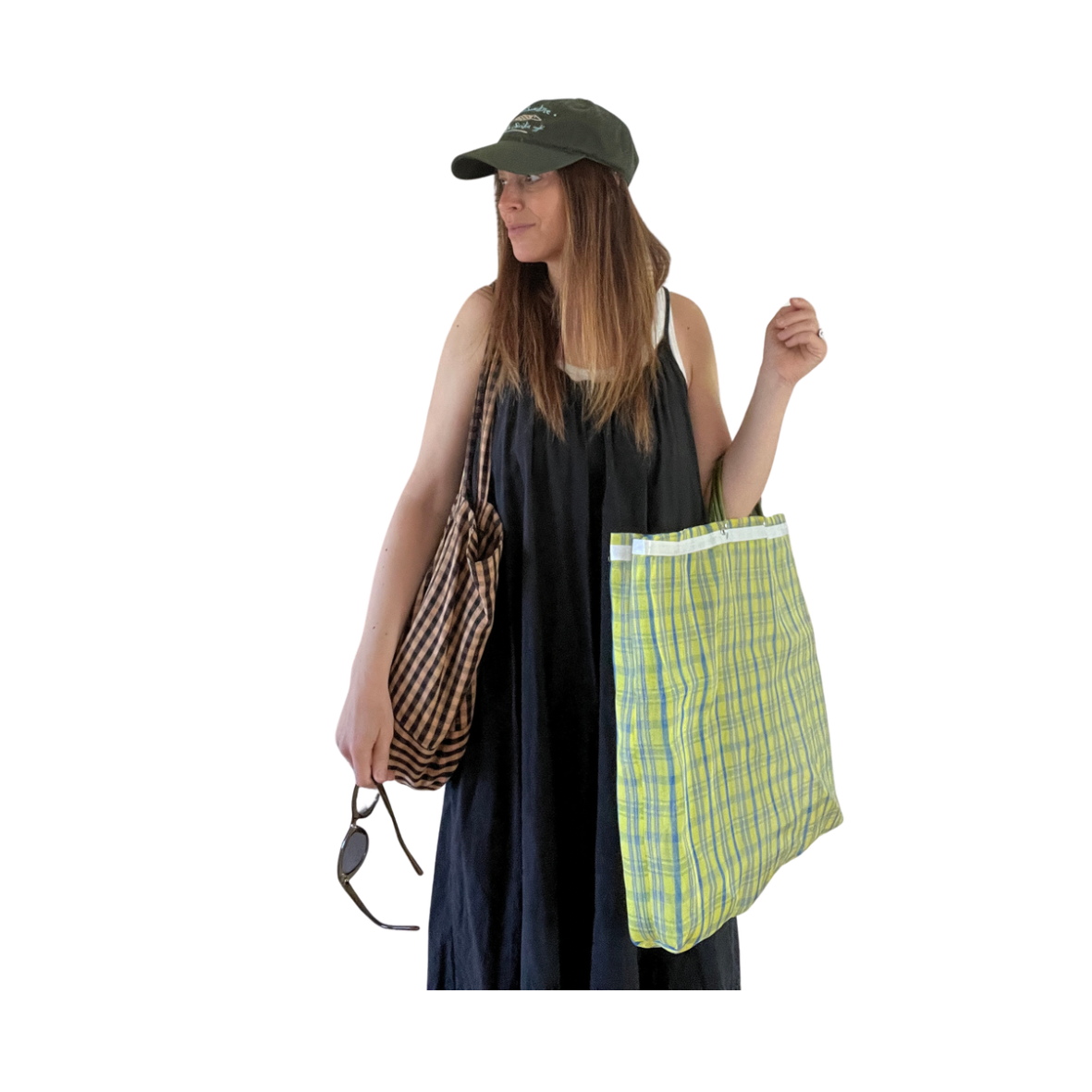 Woman dressed in black dress, wearing a green cap, holding a yellow plaid shopping bag in her right hand, and a checkered shopping bag over her left shoulder, holding a pair of sunglasses. Farmers market vibe.