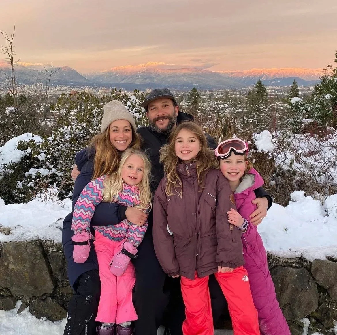 A family of five outdoors in a snowy landscape with mountains in the background during sunset, all smiling and dressed in winter clothing.
