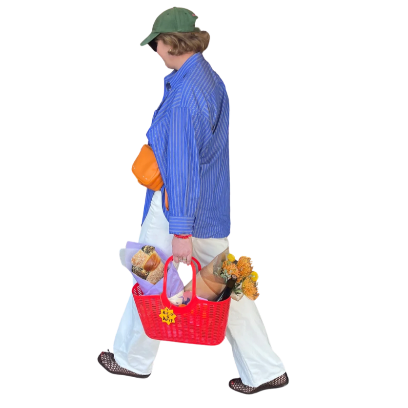 Person walking with a red shopping basket filled with groceries, wearing a green cap, blue striped shirt, white pants, and carrying a bouquet of flowers in one hand.
