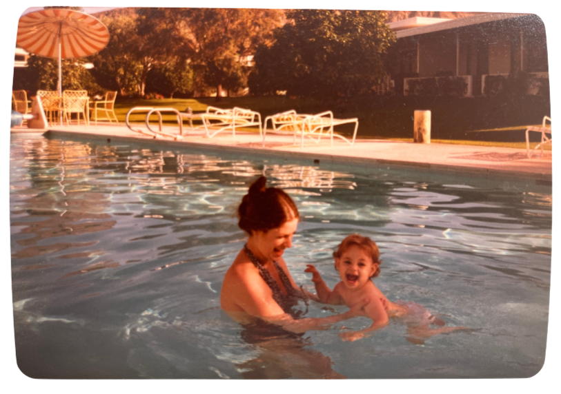 A woman and a young child enjoying a swim in an outdoor pool during sunset, with pool chairs and a large patio umbrella visible in the background.