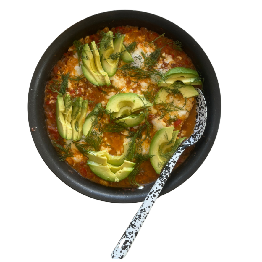 A bowl of shakshuka with avocado slices and fresh herbs and feta, served with a spoon.