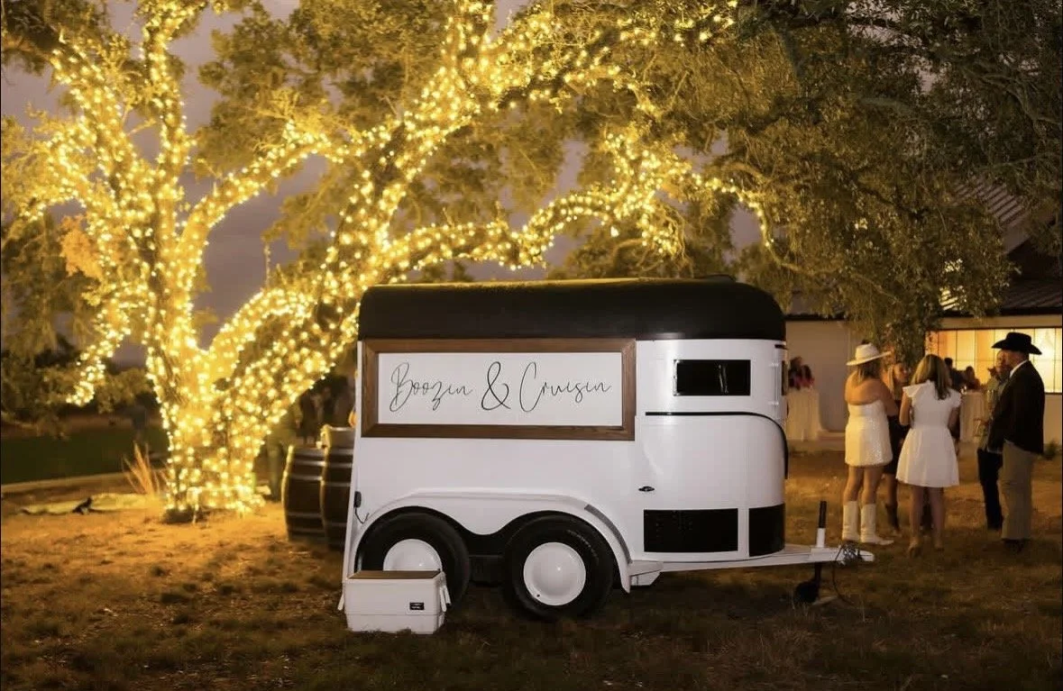 A vintage-style decorated food trailer with a sign reading 'Boozn & Crouzn' is parked outdoors at night, illuminated by a large tree wrapped in yellow fairy lights, with a group of people dressed in semi-formal attire nearby.