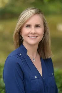 A woman with blonde hair smiling outdoors in a blue shirt, with trees and greenery in the background.