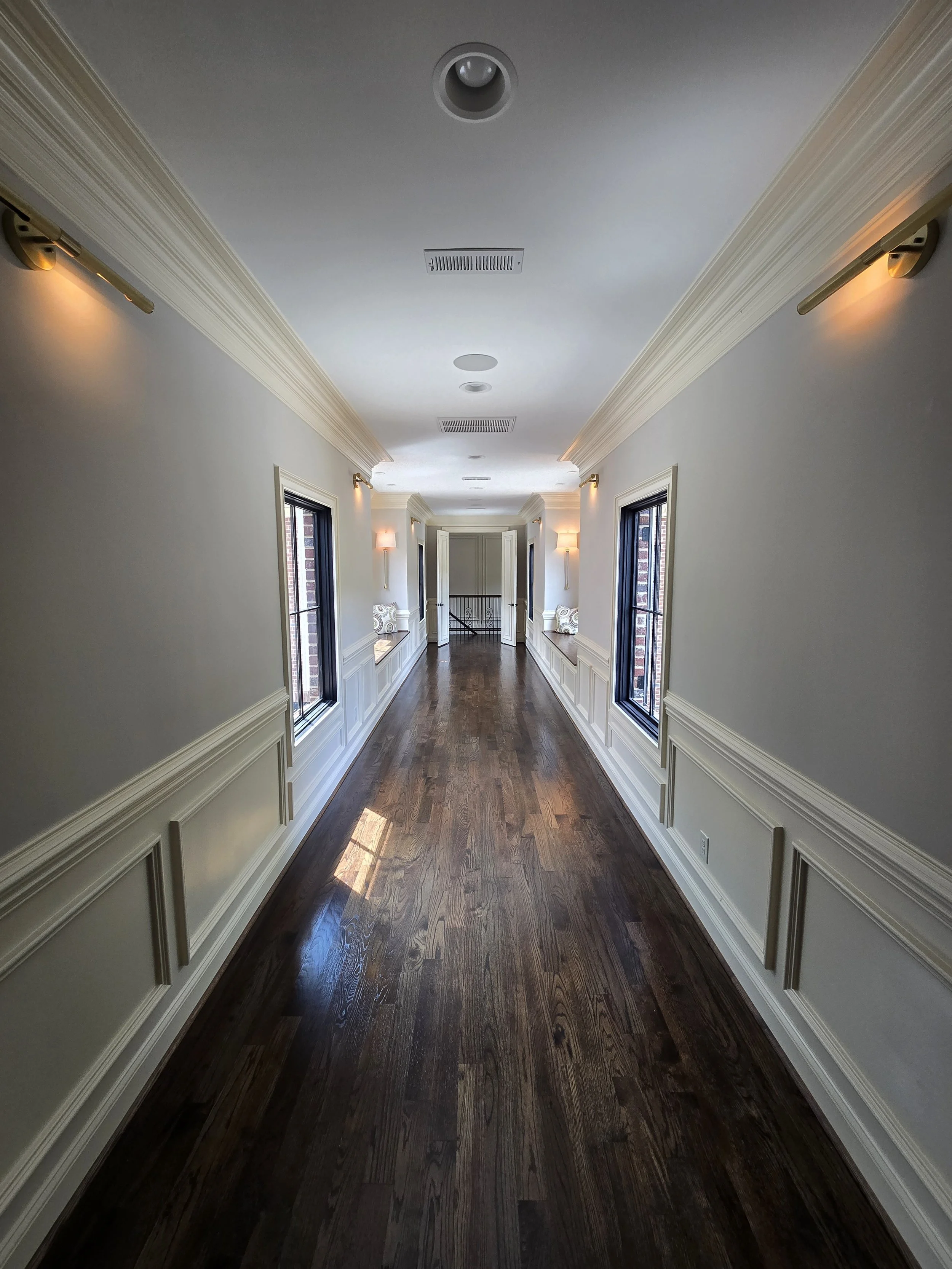 Custom home hallway by The Kiker Group in Monroe, NC, featuring dark wood flooring, decorative wall molding, black-framed windows, and elegant lighting.