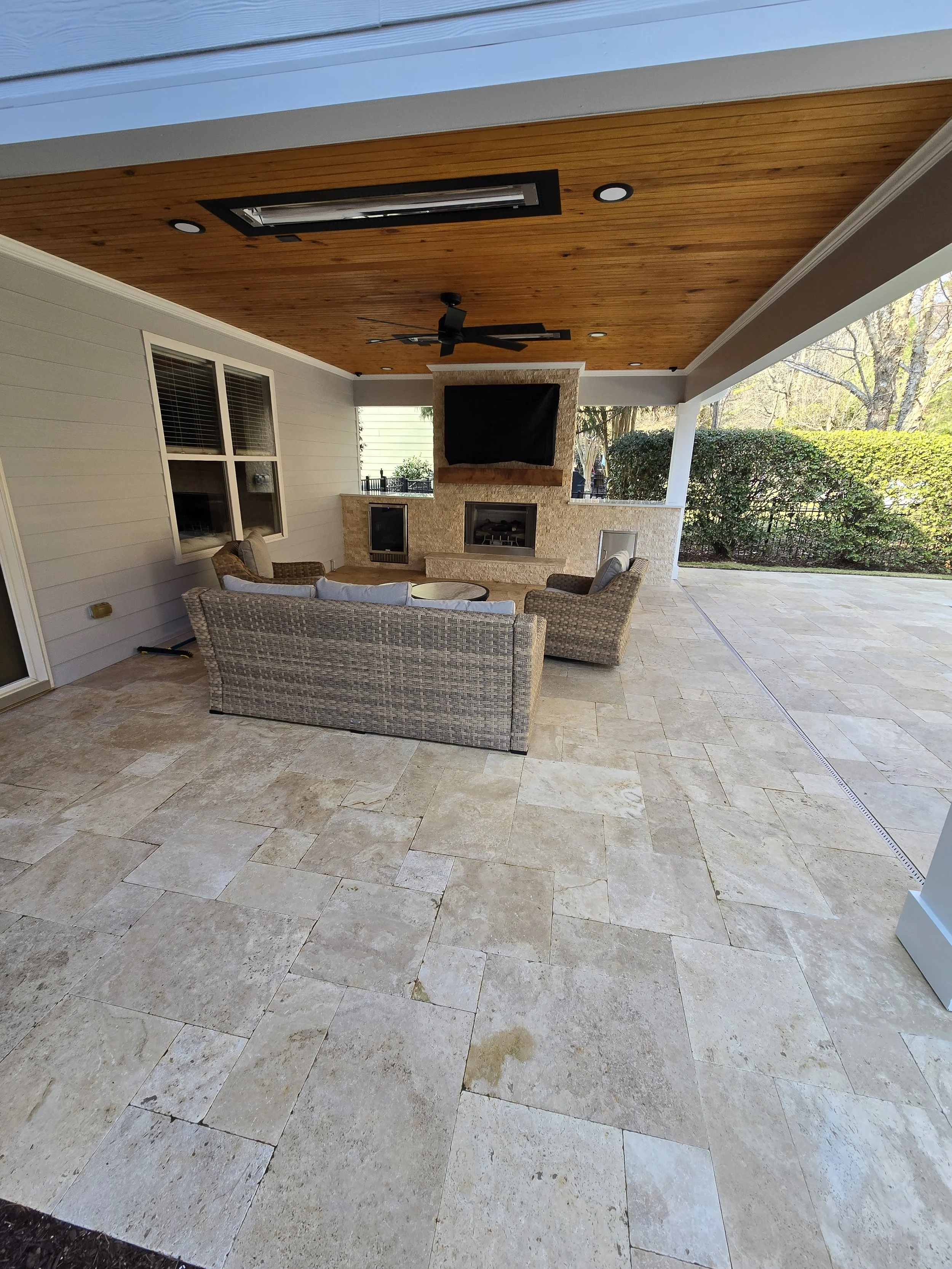 Custom covered patio by The Kiker Group in Monroe, NC, featuring stone flooring, wicker seating, an outdoor fireplace, television, and wood ceiling.