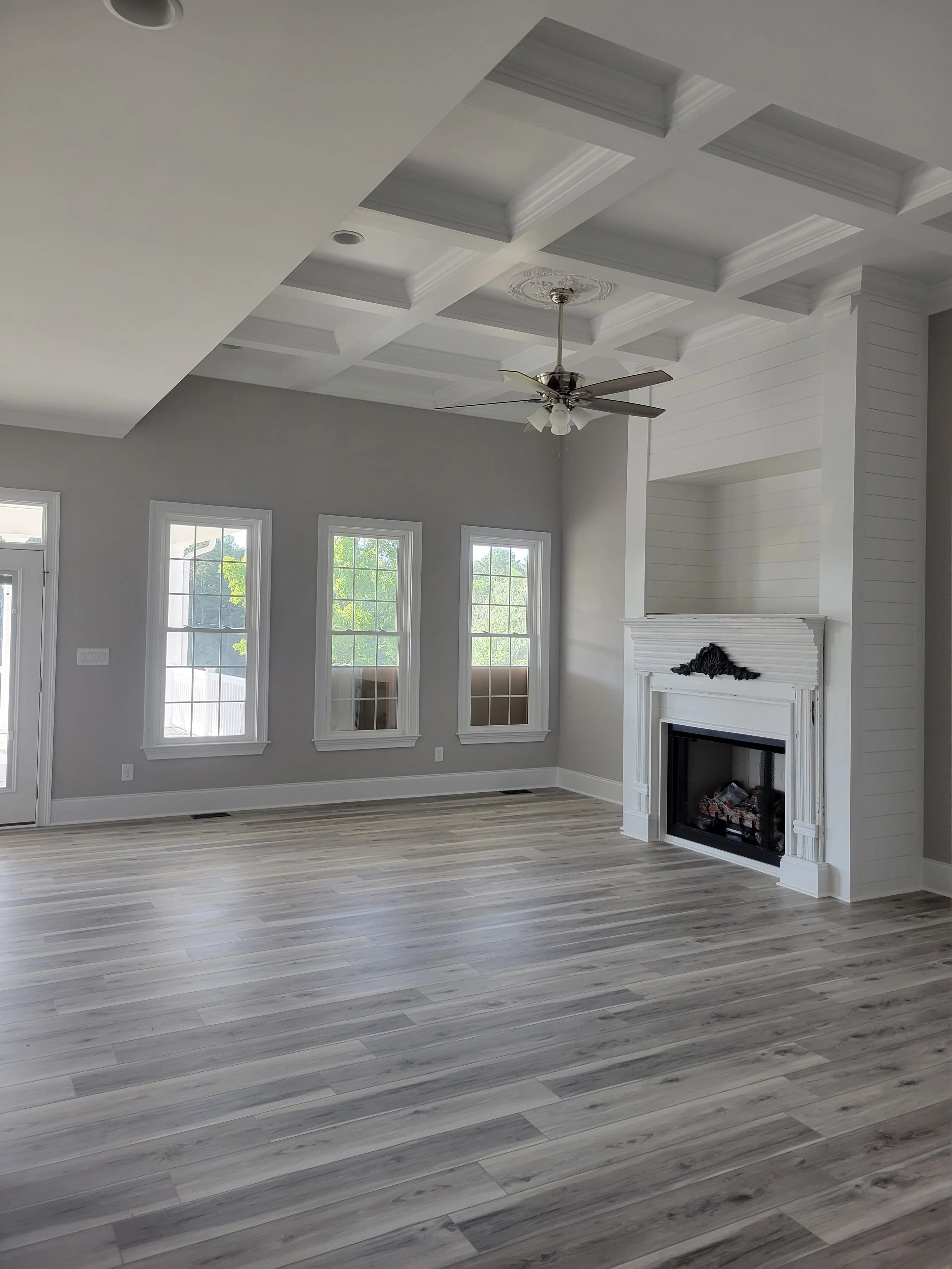 Newly constructed home interior by The Kiker Group in Monroe, NC, featuring large windows, a white fireplace, ceiling fan, and light wood flooring.
