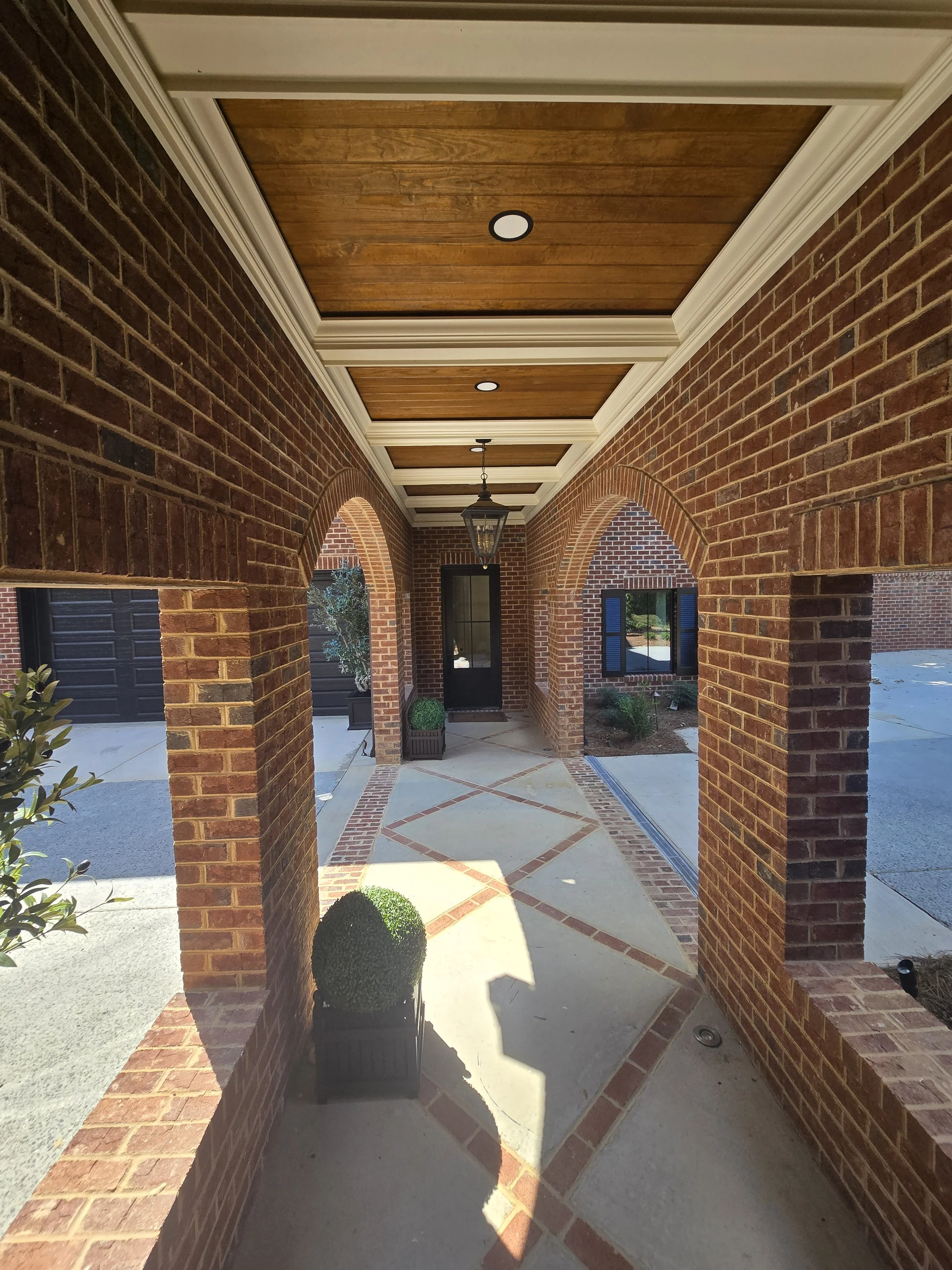 Covered brick walkway at a custom home by The Kiker Group near Monroe, NC, featuring arched openings, a wood ceiling, recessed lighting, and potted plants.