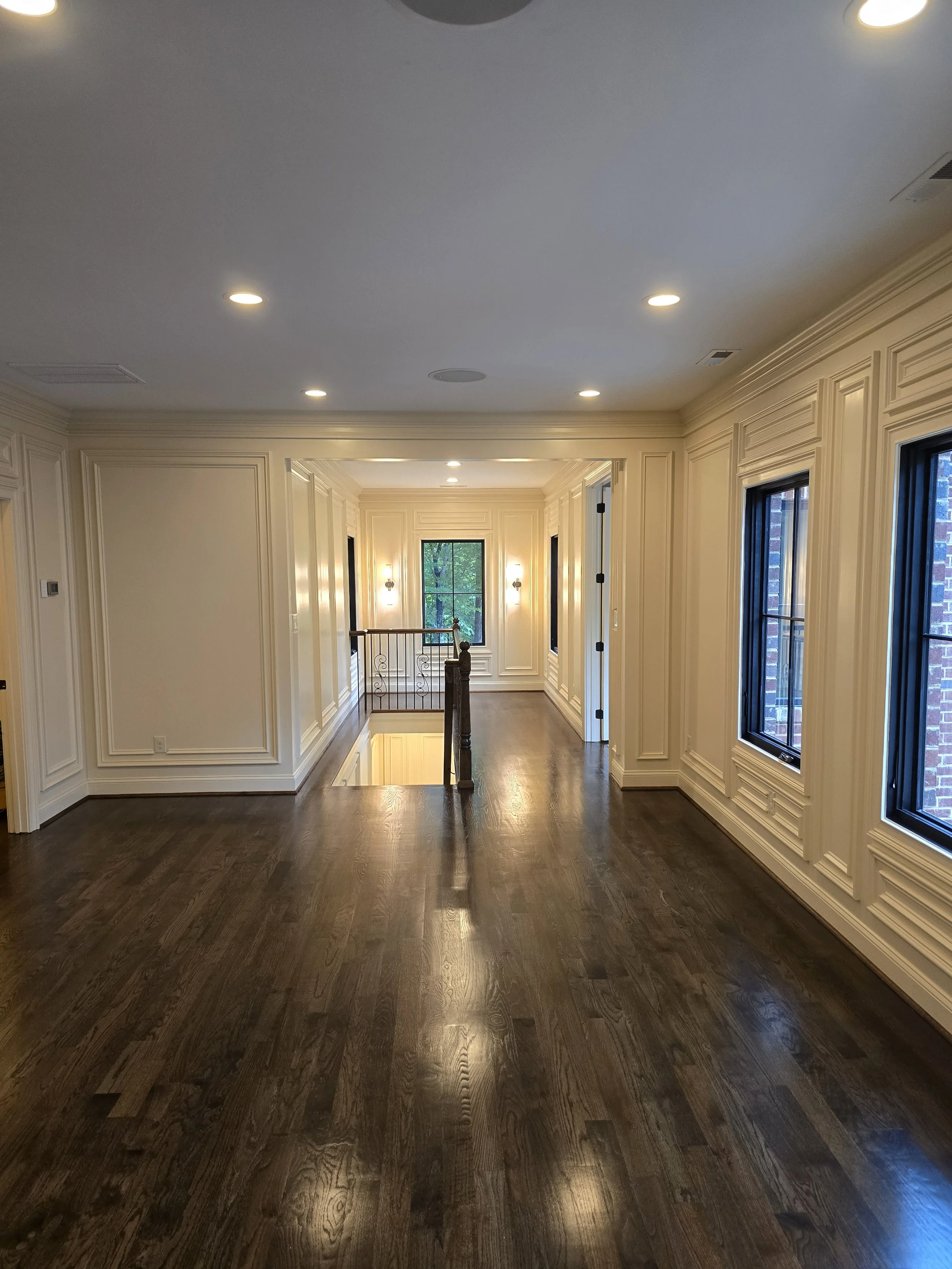 Bright custom home hallway by The Kiker Group in Monroe, NC, with dark hardwood floors, decorative molding, ceiling lights, and large windows.