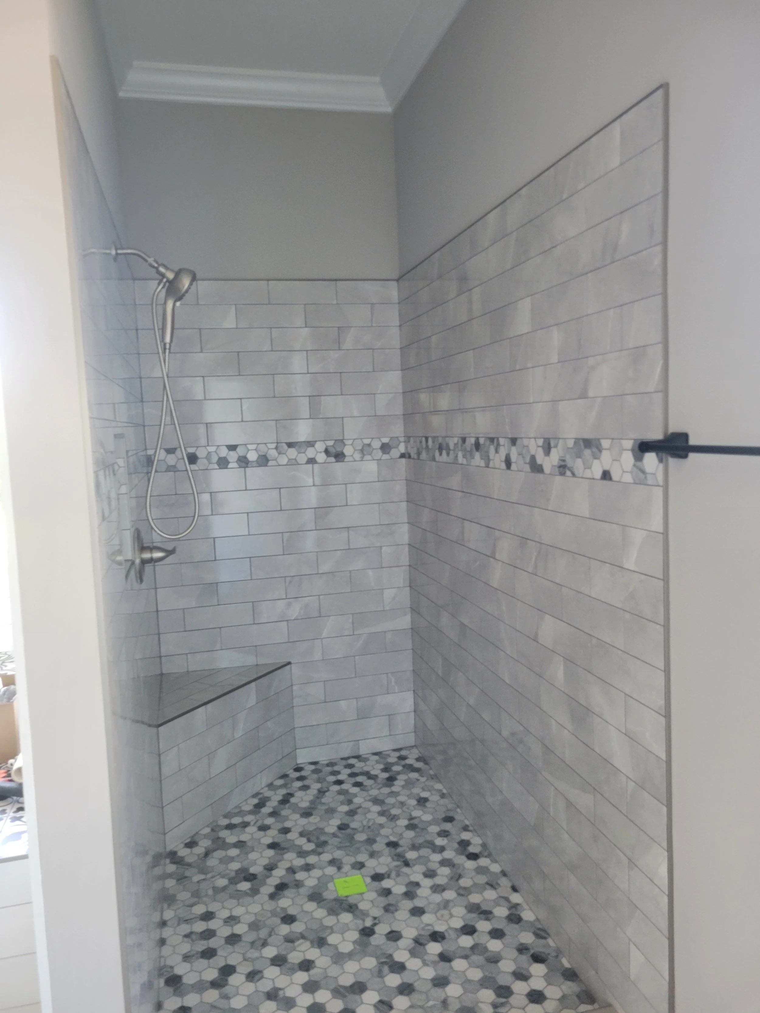 Modern custom shower by The Kiker Group in Monroe, NC, featuring gray tile walls, a decorative tile border, hexagonal flooring, and handheld showerhead.