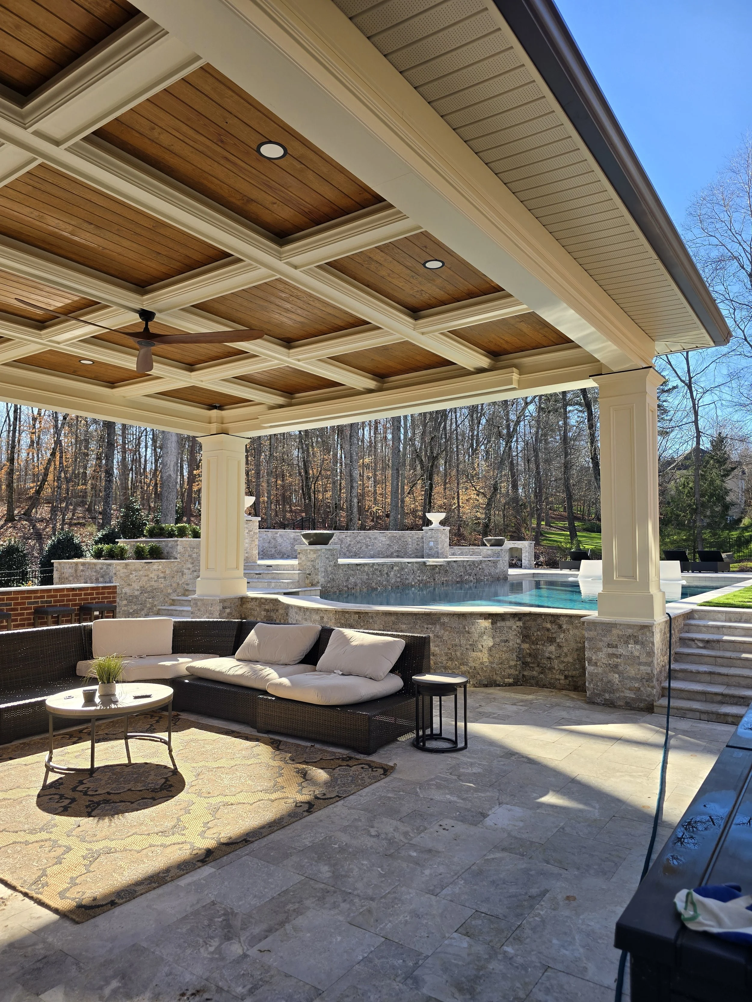Covered outdoor patio by The Kiker Group near Monroe, NC, featuring a wood ceiling, wicker seating, ceiling fan, and views of a custom pool with a stone fountain.