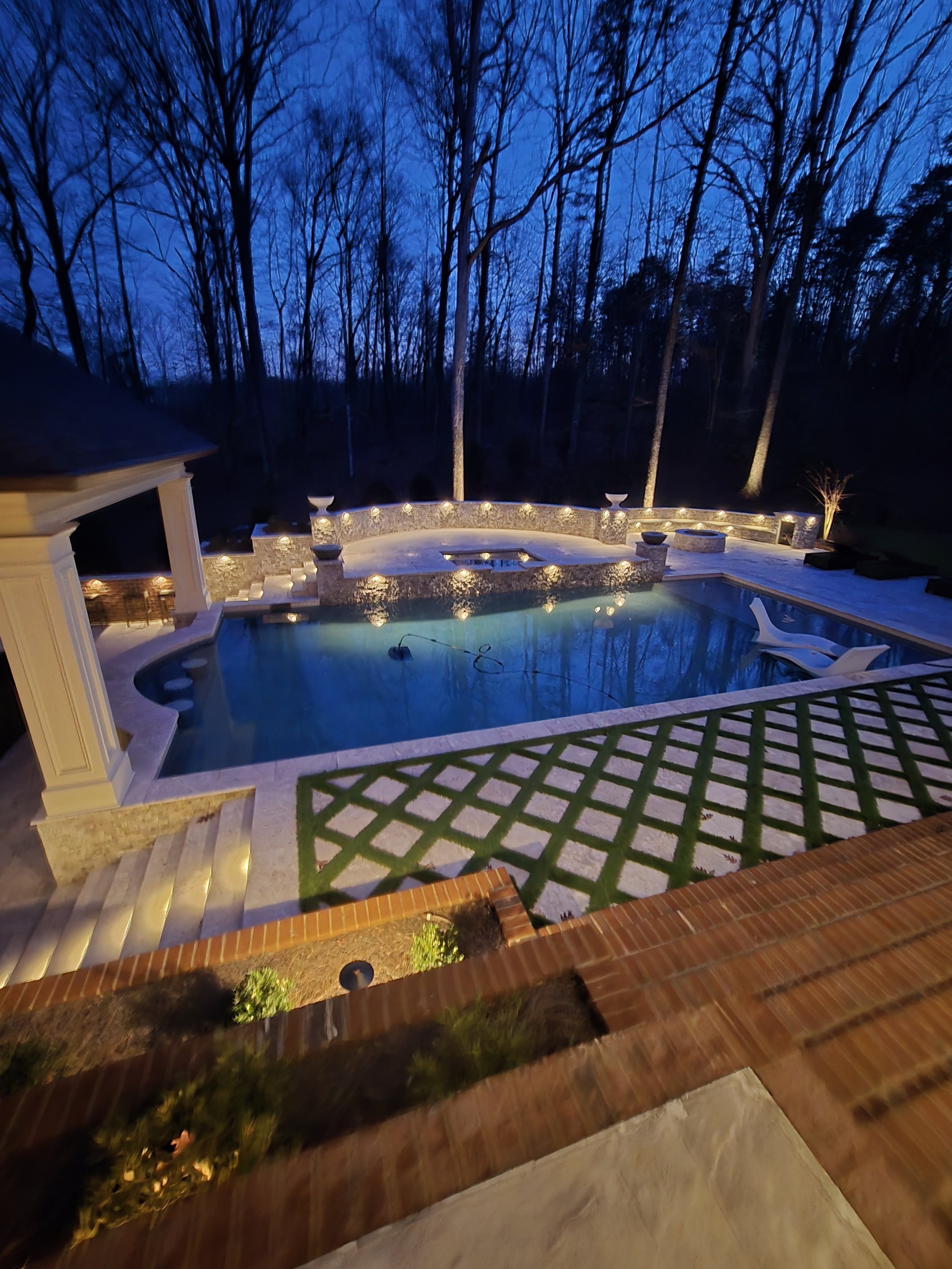 Custom backyard swimming pool by The Kiker Group near Monroe, NC, surrounded by a stone patio and outdoor lighting at dusk.