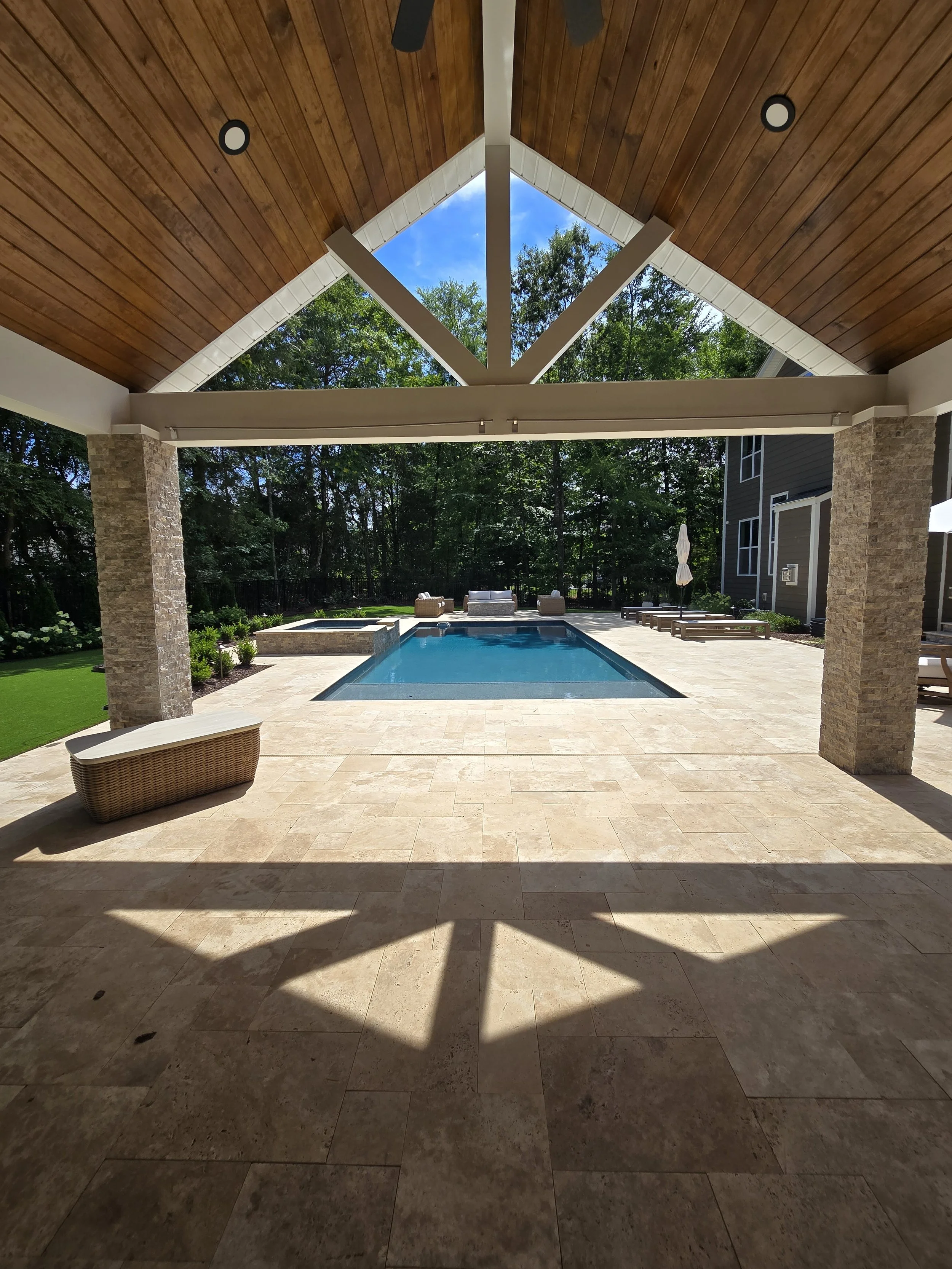 Outdoor patio area with a swimming pool, surrounded by lounge chairs and seating, under a wooden ceiling with ceiling fans and a view of trees and blue sky.