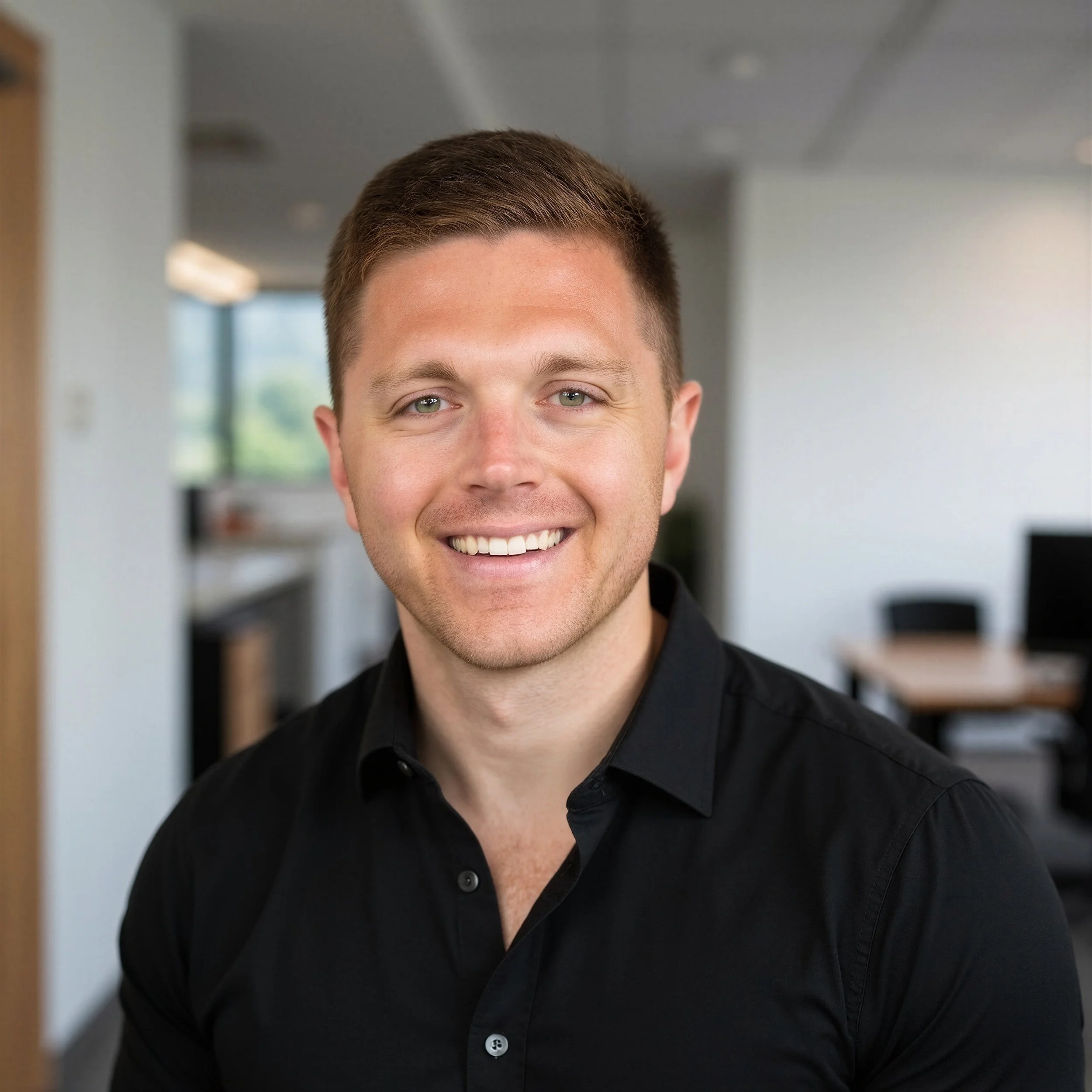 Close-up of a man with short brown hair, hazel eyes, and a wide smile, wearing a black shirt, standing indoors with blurred background.