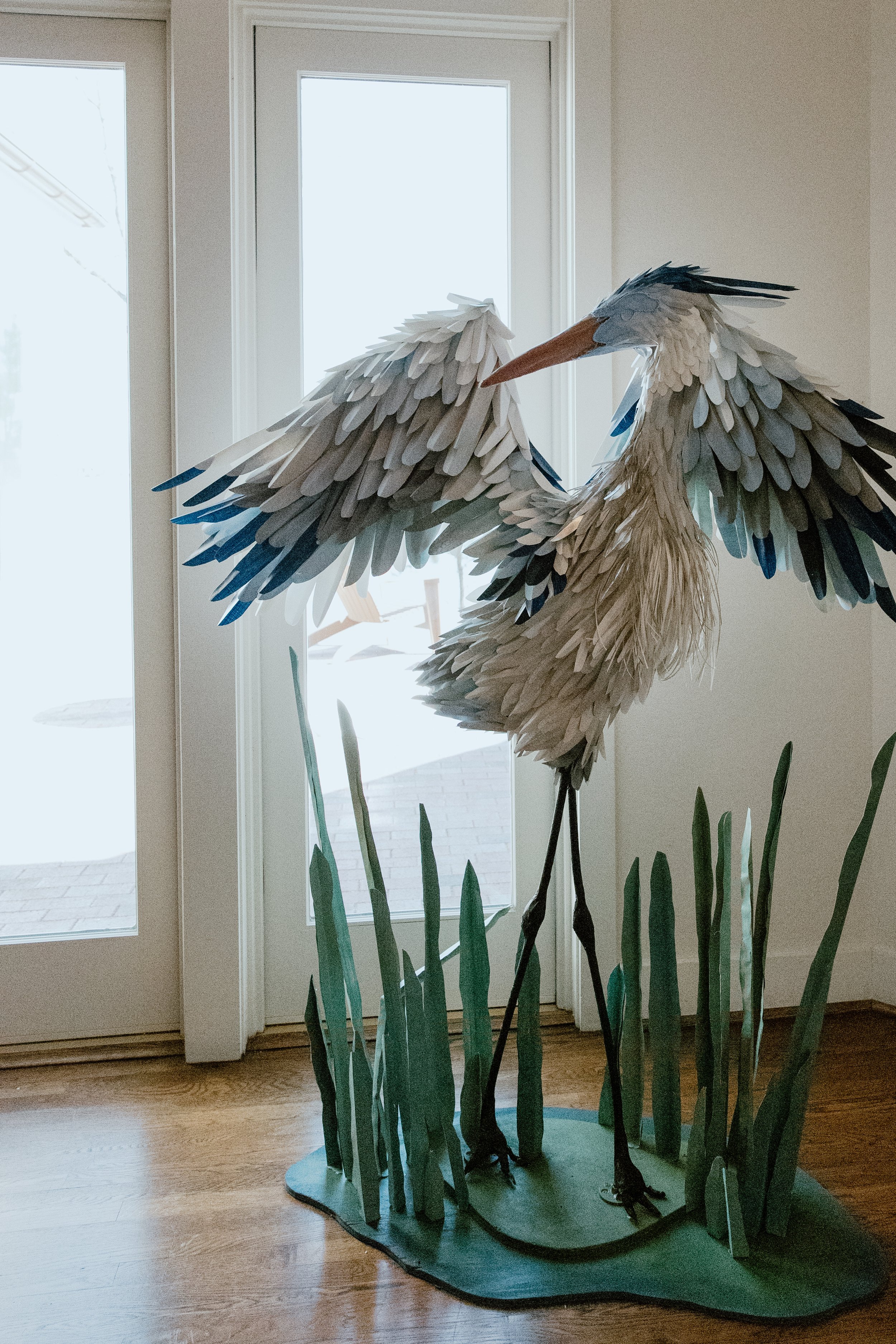 A large sculpture of a heron made out of paper or cardboard stands inside near a glass door. Tall green paper or cardboard grass surrounds the heron at its base.