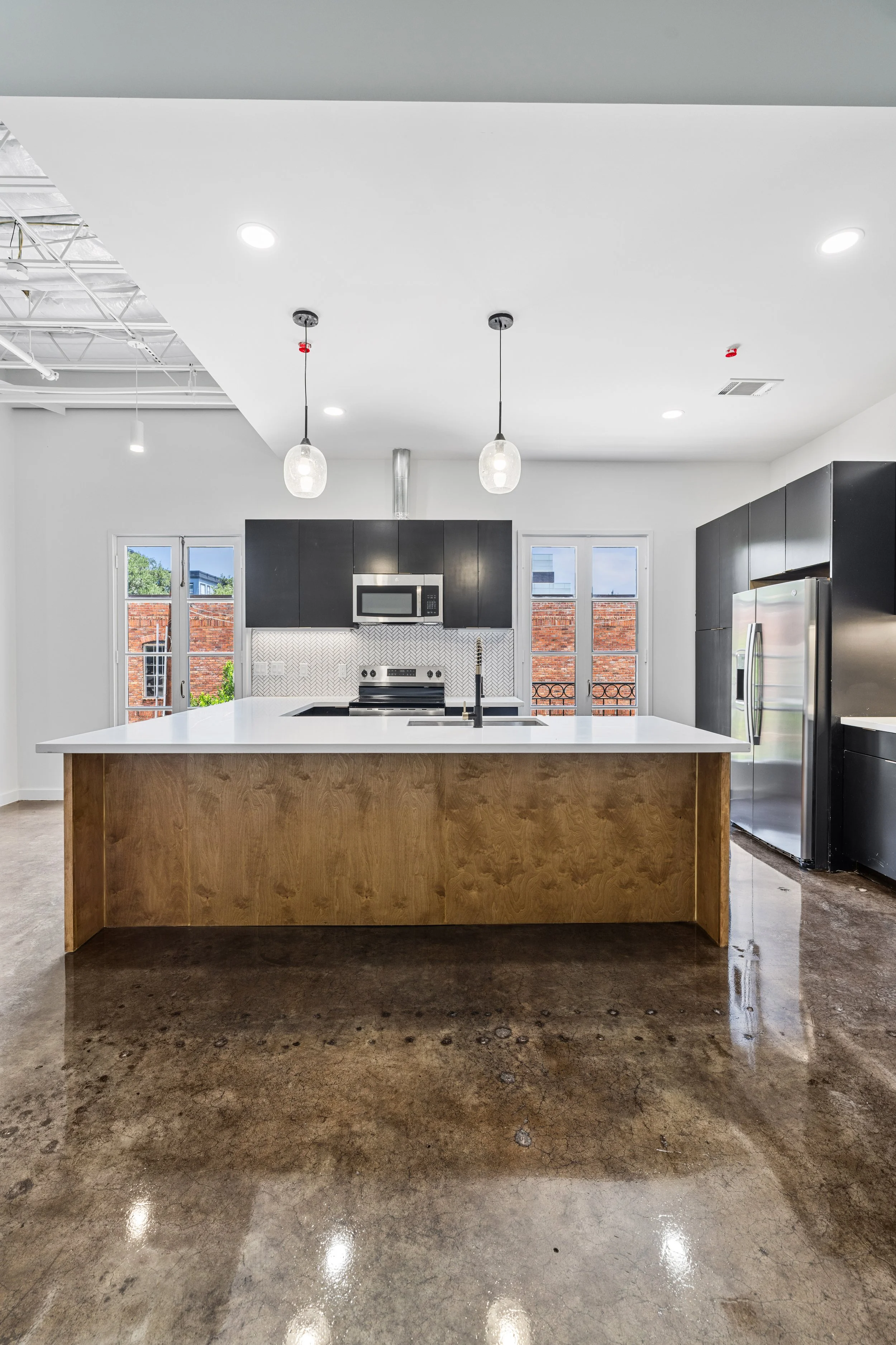 Modern kitchen with white island, dark cabinets, stainless steel appliances, large windows, pendant lights, and polished concrete floors.