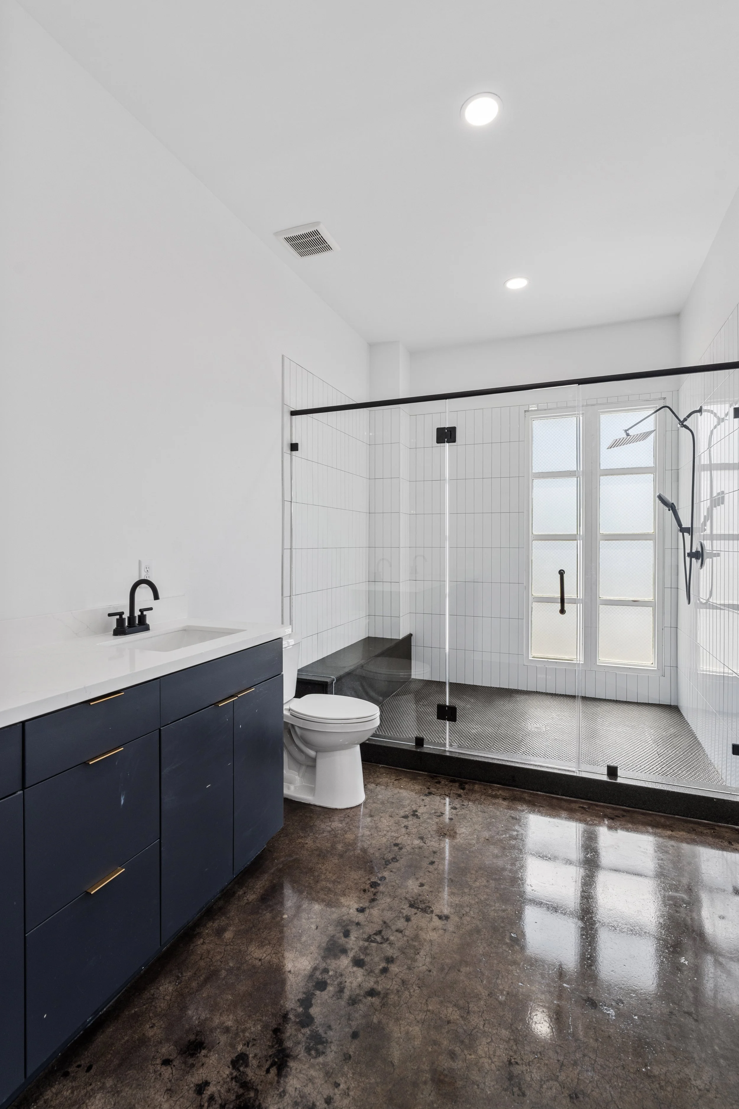 Modern bathroom with dark blue vanity, white sink, black faucet, toilet, glass shower with black frame, white tile walls, large window, and polished concrete floor.