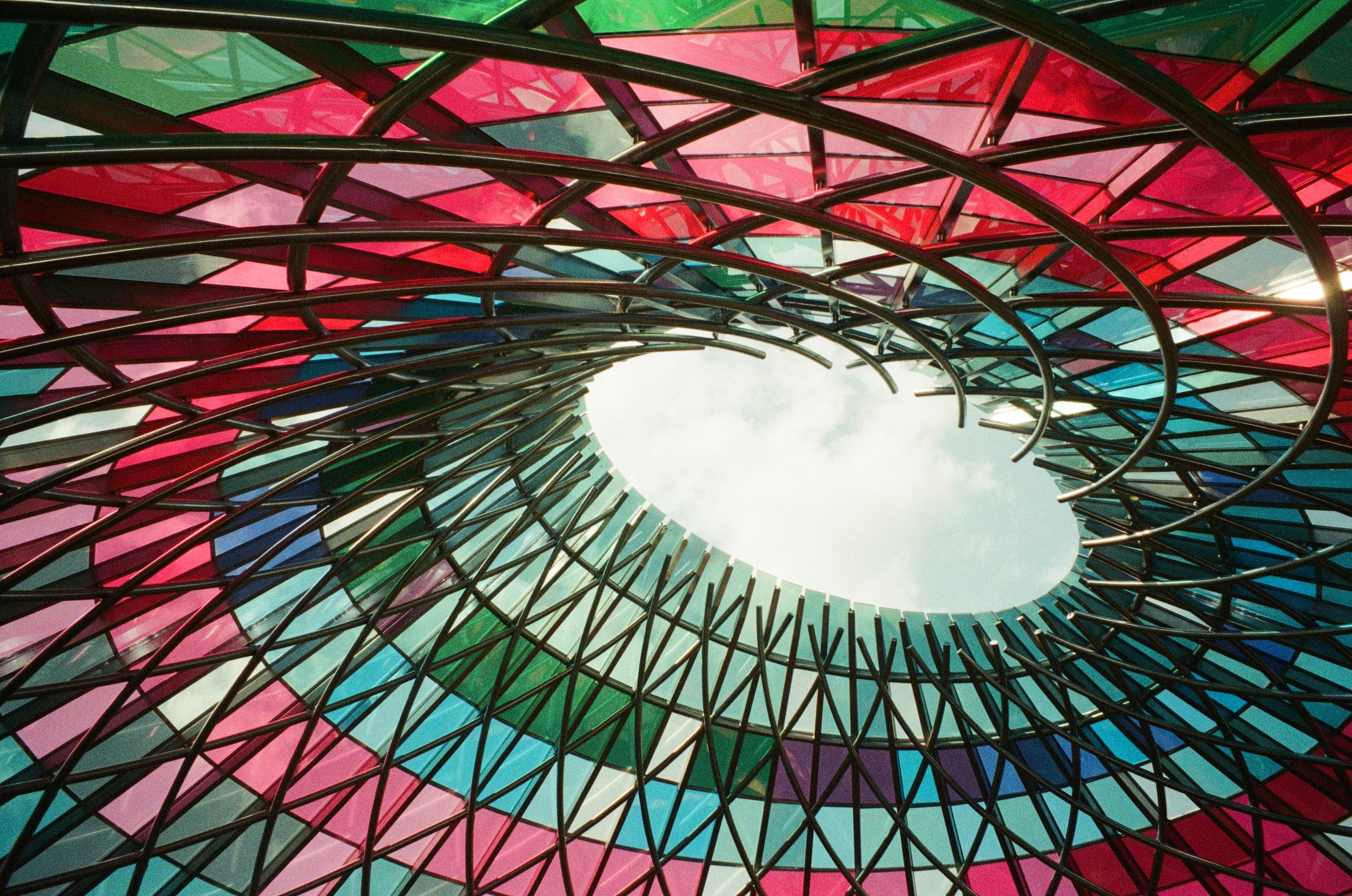 Close-up view of an architectural structure composed of multicolored glass panels in pink, red, blue, and green, held together by black metal framework, forming a tunnel-like shape with an opening to the sky visible at the center.