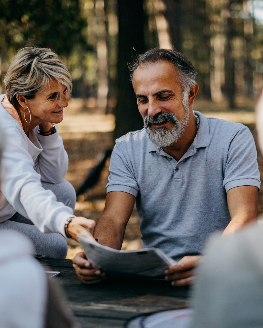 A middle-aged man with gray hair and a beard, wearing a light blue polo shirt, looks at a photo or document held by a woman with short blonde hair and hoop earrings, in a wooded outdoor setting with sunlight filtering through trees.