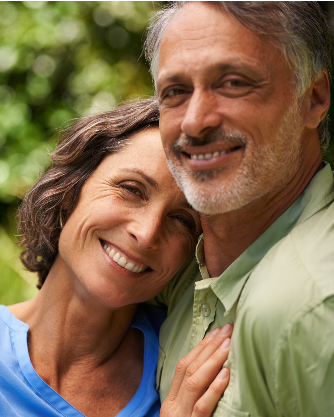 Close-up of a smiling middle-aged couple outdoors with green foliage in the background, embracing each other.