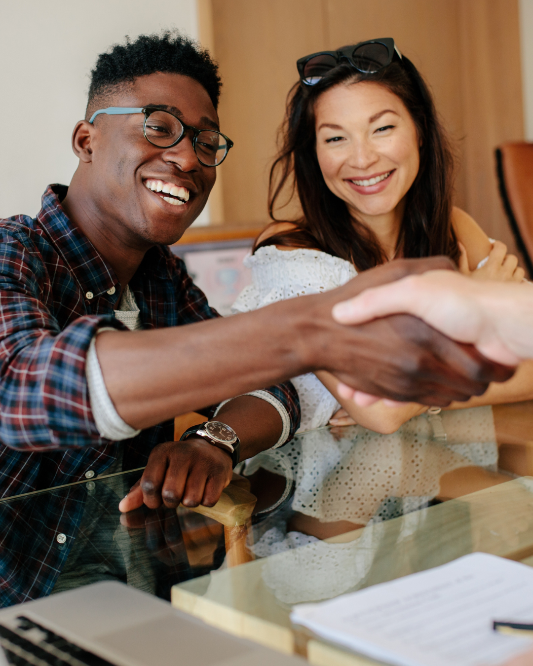 Two smiling young adults shaking hands at a meeting table with documents and a laptop.