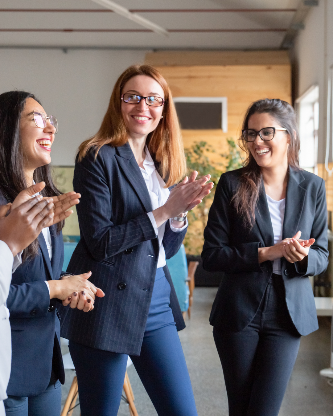 Group of women in business attire smiling, clapping, and celebrating in a modern office space.