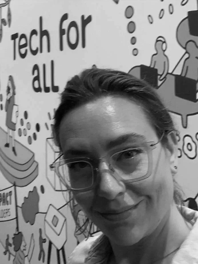 A woman with glasses taking a selfie in front of a wall with a tech-themed mural that includes illustrations of people, computers, and the words "Tech for all."