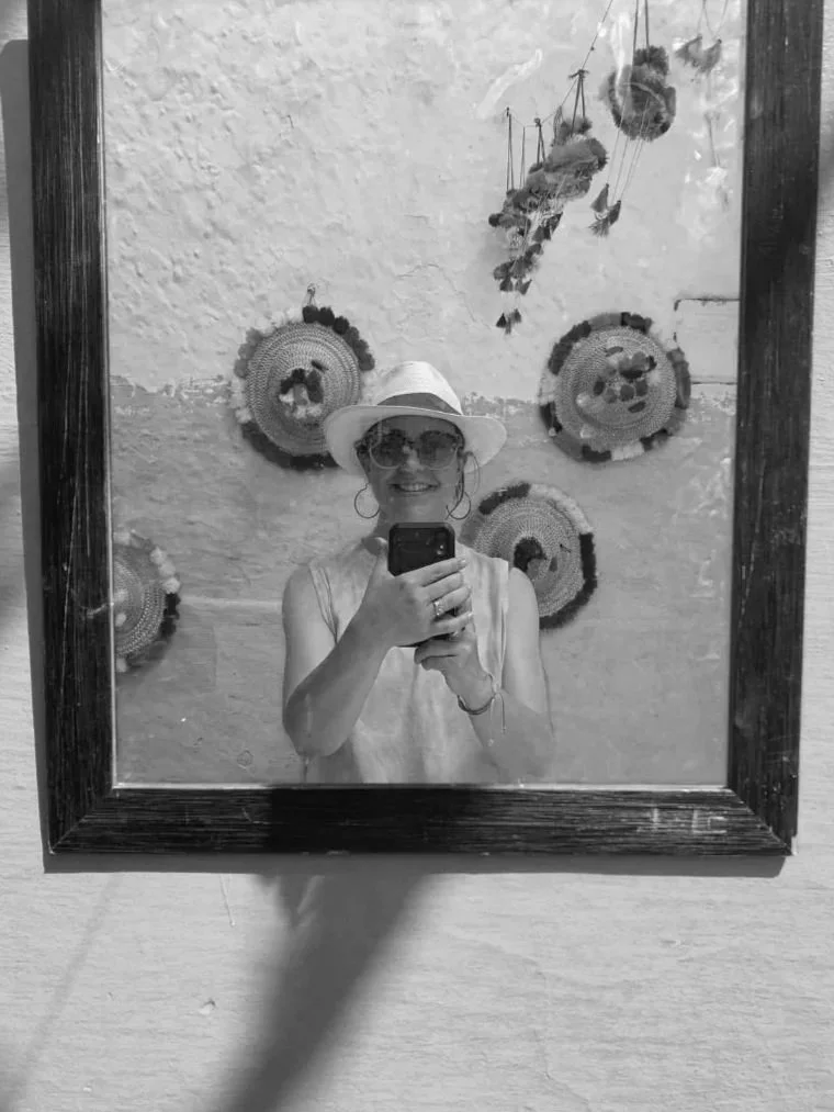 A woman taking a selfie in a mirror, wearing sunglasses, a hat, and earrings. Behind her, there are decorative circular wall hangings and hanging plants. The photo is in black and white.