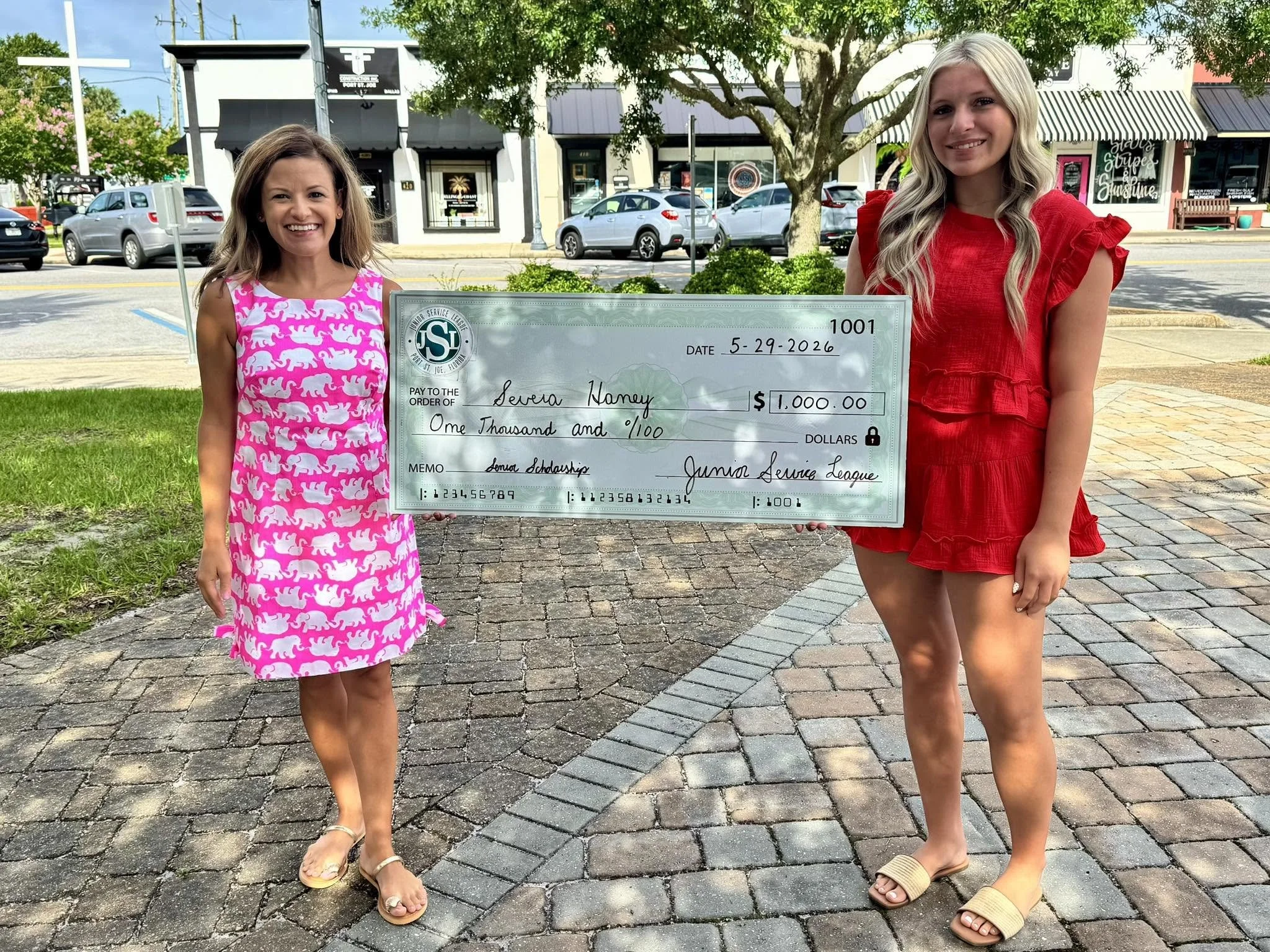 Two women standing outdoors on a brick sidewalk and holding a large check for $1,000. The woman on the left is wearing a pink and white dress with a pattern of elephants and sandals, while the woman on the right is dressed in a red dress with ruffled sleeves and beige slides. There are trees, parked cars, and storefronts in the background.