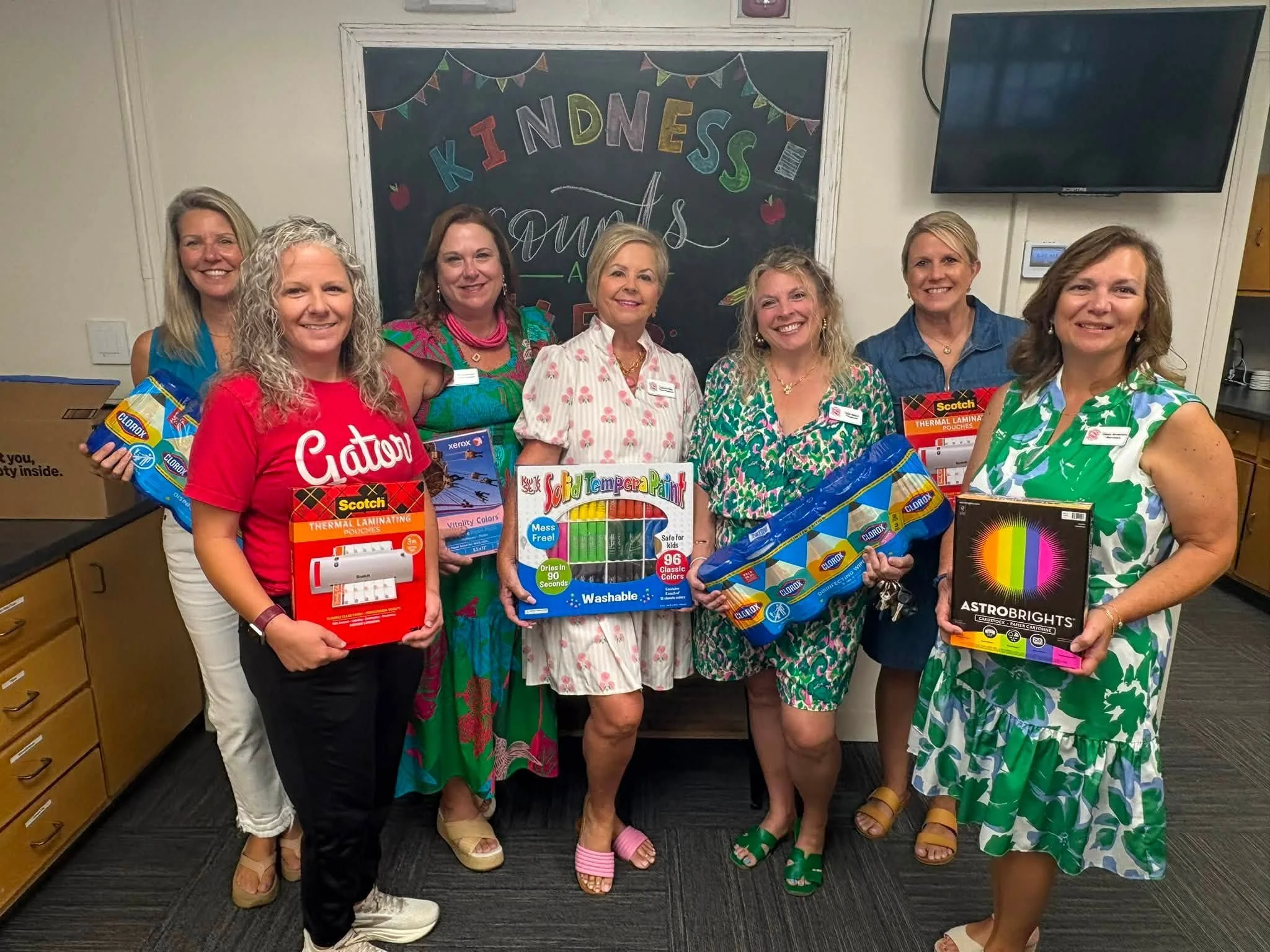 Group of women holding various school supplies, standing in front of a chalkboard with colorful letters spelling "KINDNESS" and "SQUAD" in a classroom or office setting.