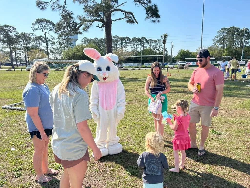 A group of people, including children and adults, are outdoors at an event with a person in a white Easter Bunny costume. The scene is in a grassy park with trees and a tennis court in the background. Two children are interacting with the Bunny, and others are standing nearby.