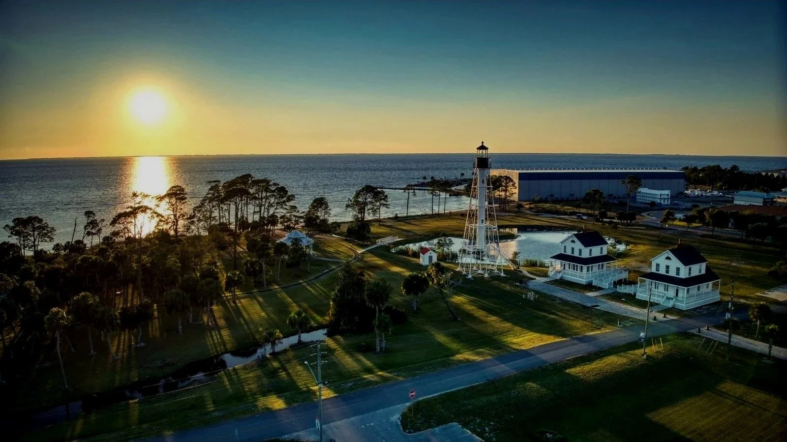 Sunset over a coastal park with a lighthouse, small white houses, trees, a pond, and a large building in the background.