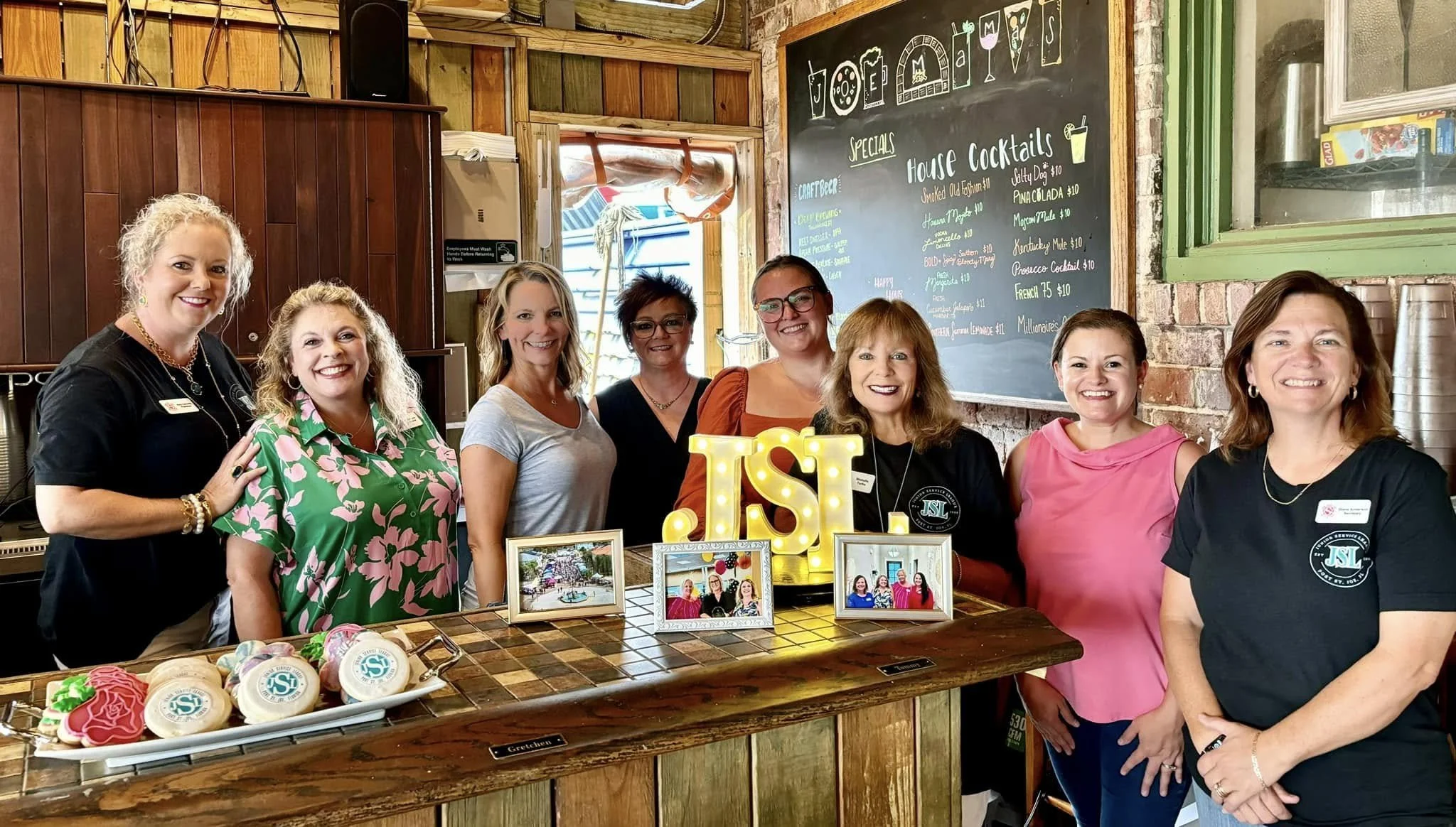 Group of women smiling for a photo at a Junior Service League New Member gathering, standing behind a table with decorated cookies and framed photographs, with a chalkboard menu and decorative lit sign in the background.