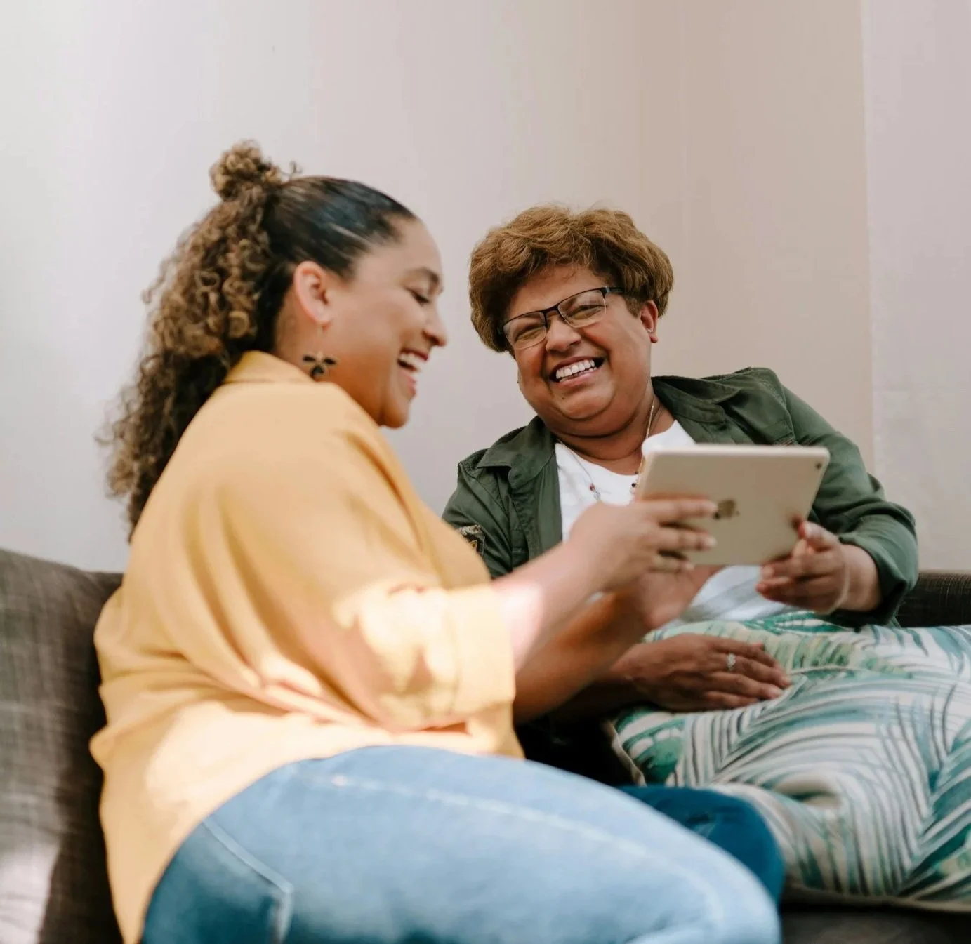 Two women sitting on a couch, sharing a laugh as they look at a tablet together.