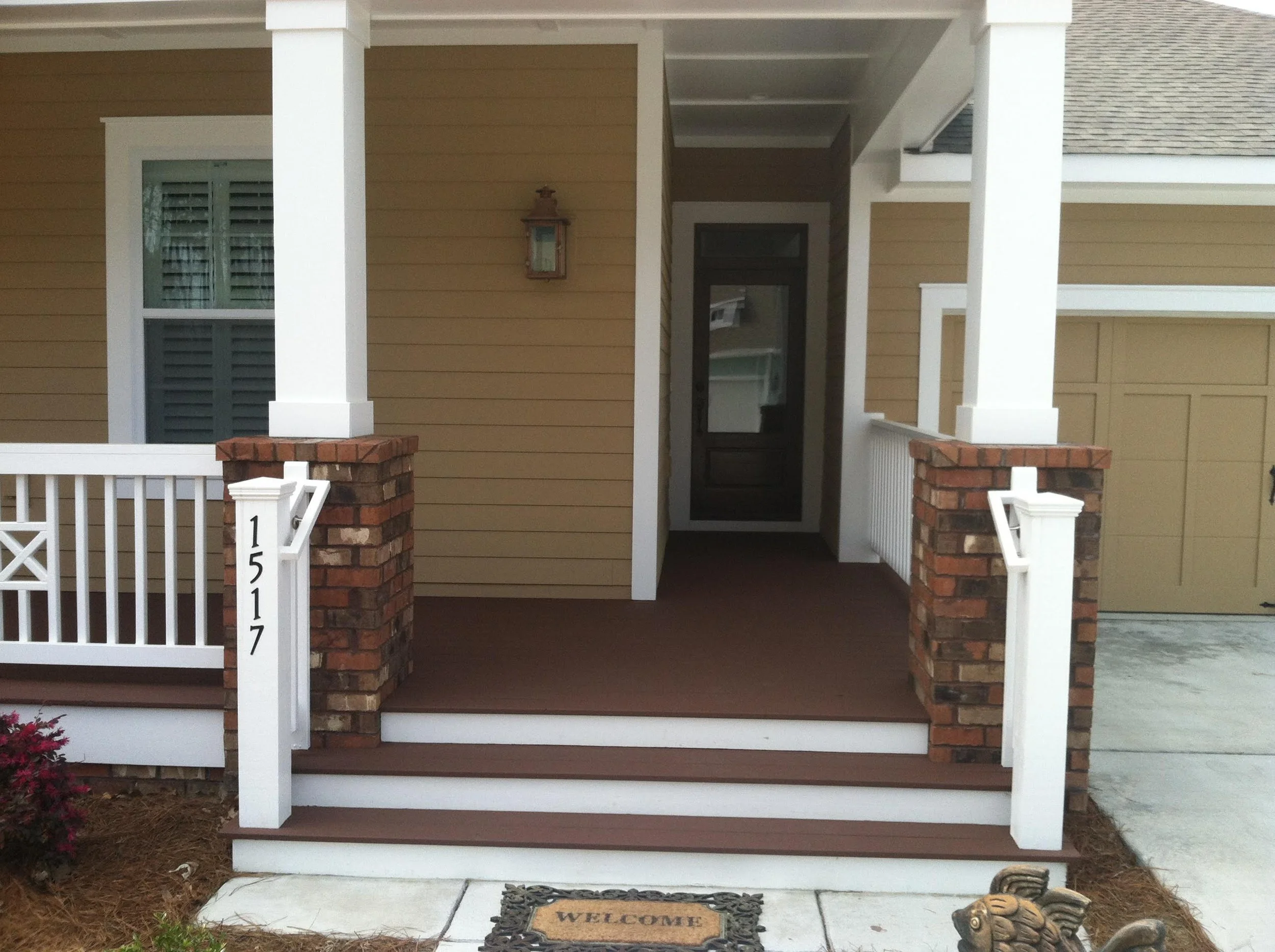 Front porch of a house with beige siding, white trim, brick pillars, a white railing, and a welcome mat with the word 'welcome' in front of the door.
