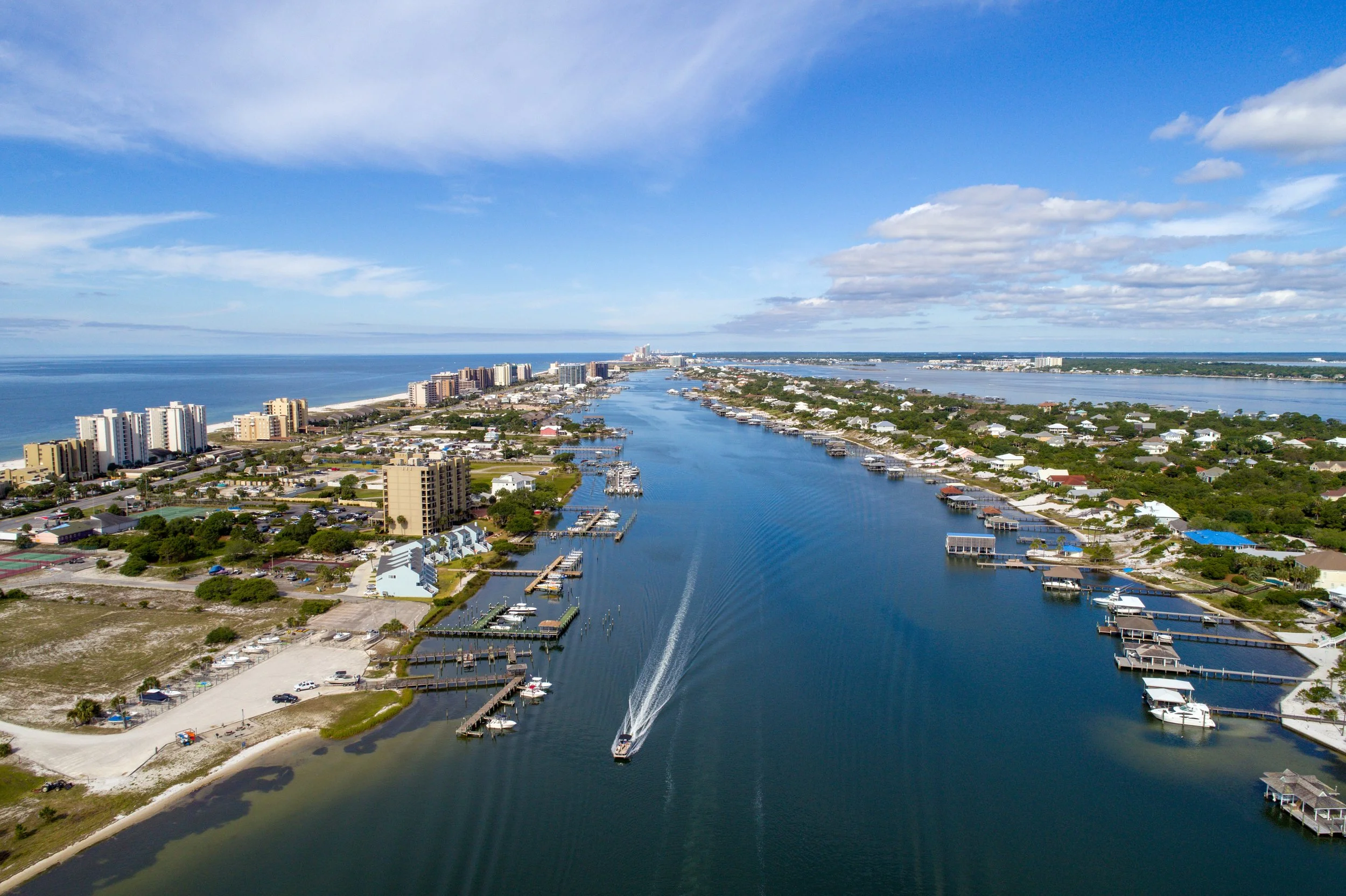 An aerial view of a coastal city with buildings, marinas, and waterways under a partly cloudy sky.