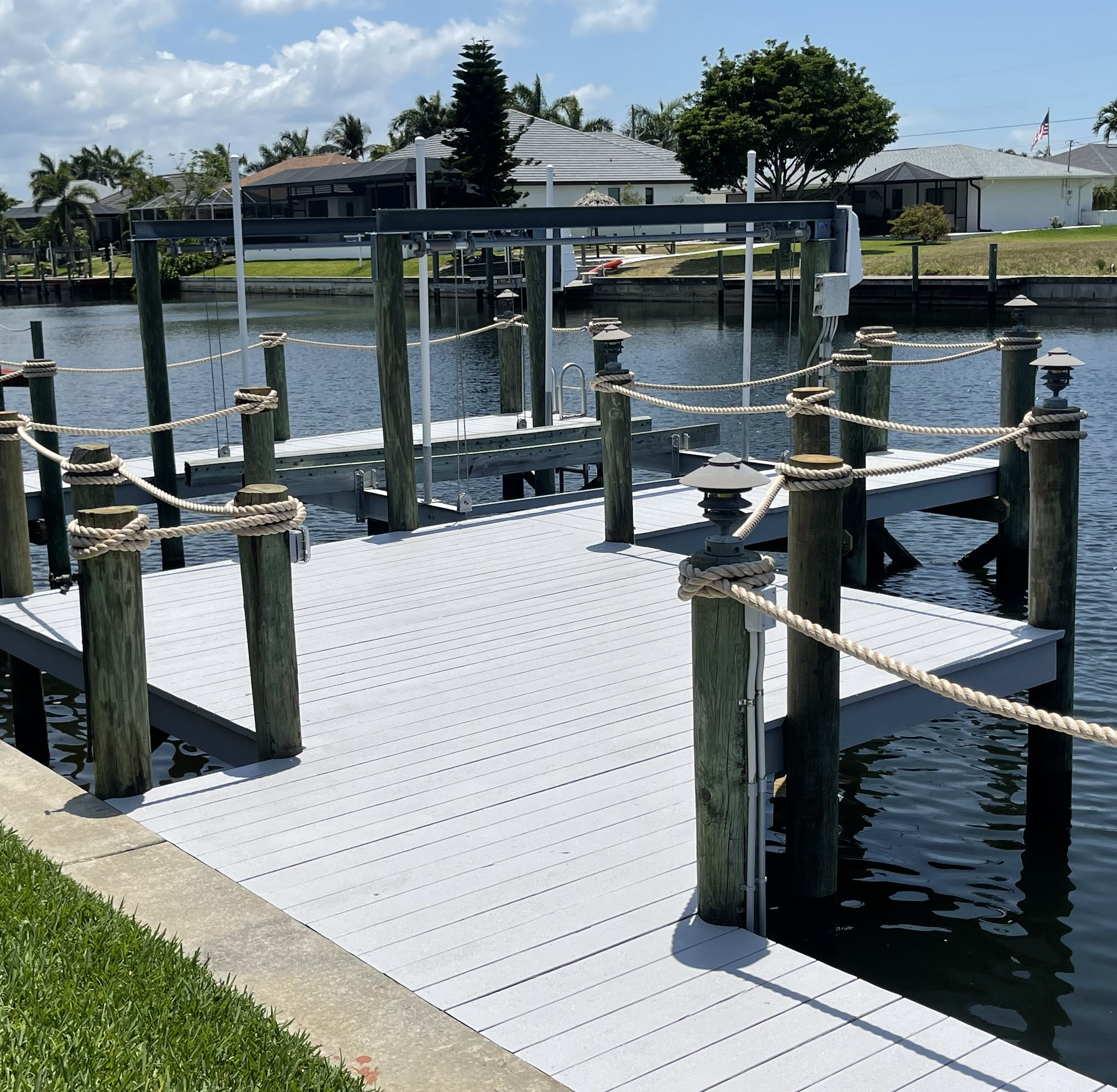 View of a backyard waterfront area with a dock, boat lift, and modern houses in the background on a sunny day with some clouds.
