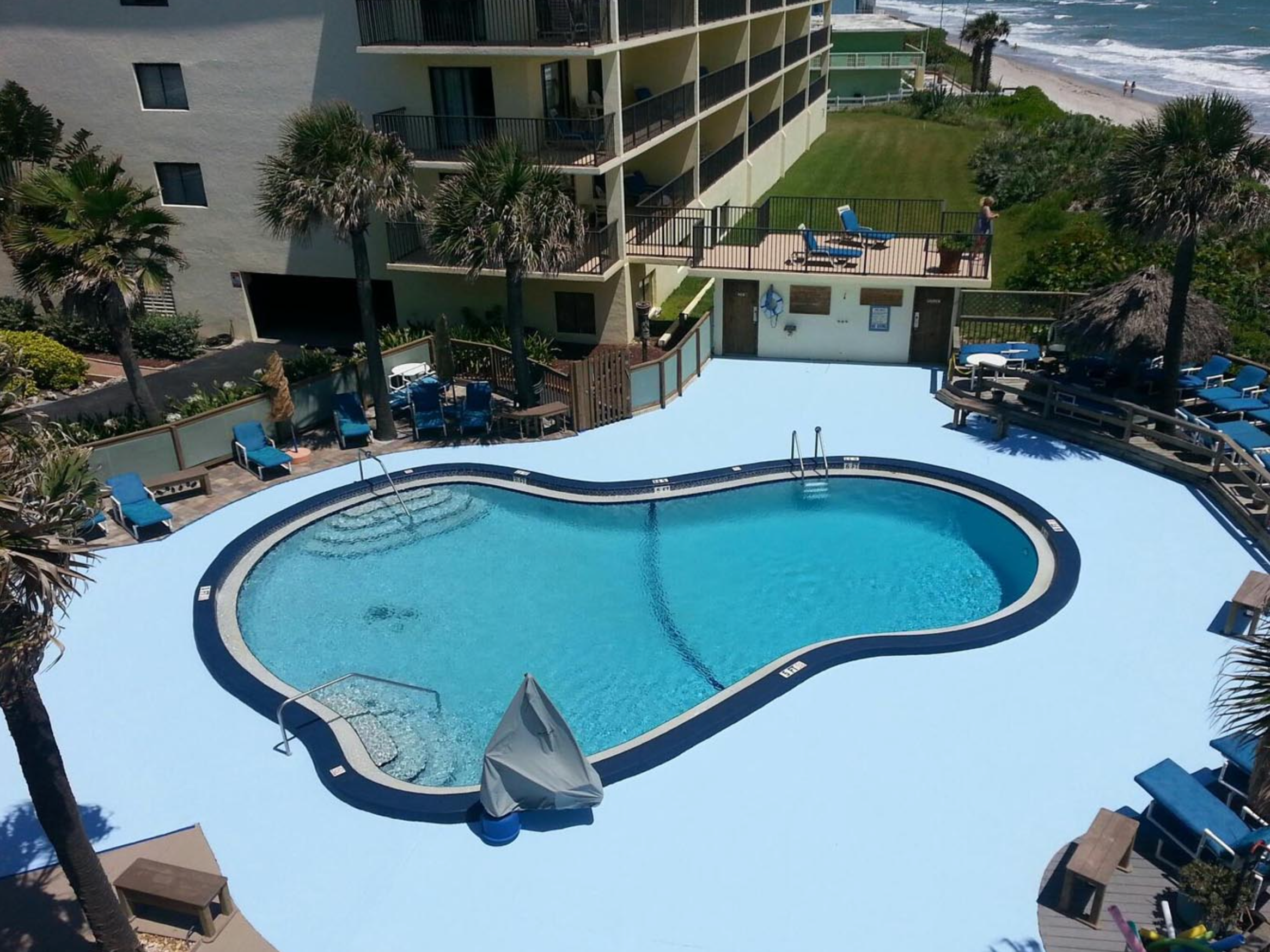 View of a hotel or apartment complex with a swimming pool and a view of the beach and ocean in the background. There are lounge chairs, umbrellas, and palm trees around the pool area.