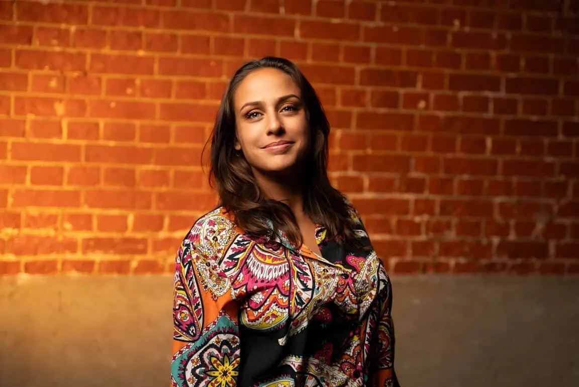 Portrait of Laila Mckenzie with long wavy dark hair wearing a colorful patterned top, standing against a warmly lit orange brick wall.