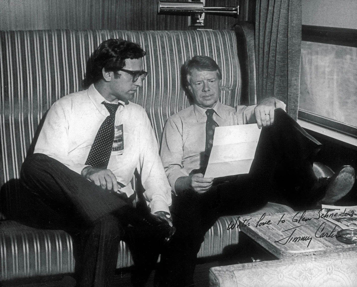 Two men in dress shirts and ties sitting on a striped bench seat on a train. One man is reading a newspaper, and the other man is looking at it. One man is wearing glasses and holding a folder on his lap. The train is moving past a window. There is a handwritten message at the bottom that says, "With love to Glen Schmendes, Jimmy Carter."