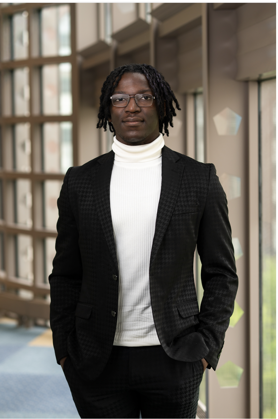 A young Black man with glasses and dreadlocks, wearing a black patterned suit and white turtleneck, standing indoors with large windows and a modern building background.