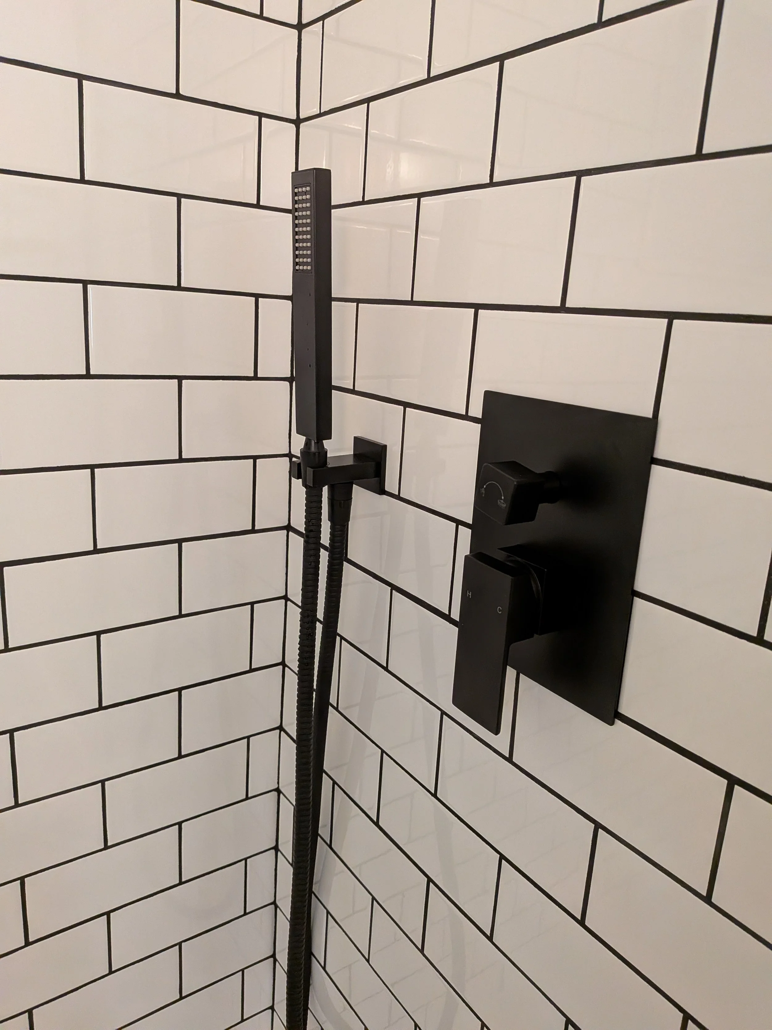 A shower with a black handheld showerhead mounted on a black hose, attached to a black wall fixture, against white subway tiles with black grout.